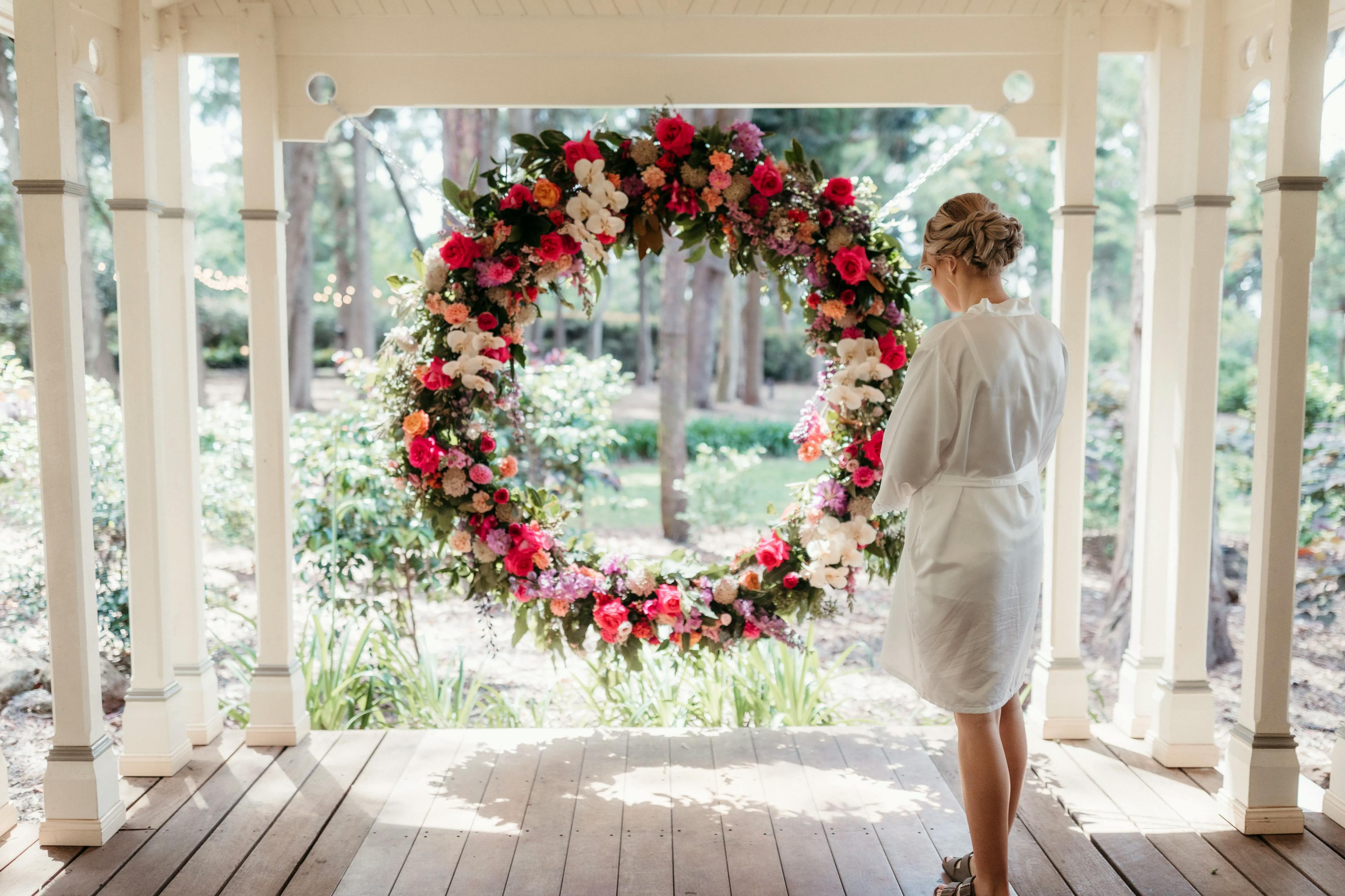 A woman in a white robe stands on a wooden veranda, looking at a large circular floral arrangement hanging in the center. The arrangement features a variety of colorful flowers, including pink, red, and white, with greenery. A garden is visible in the background.