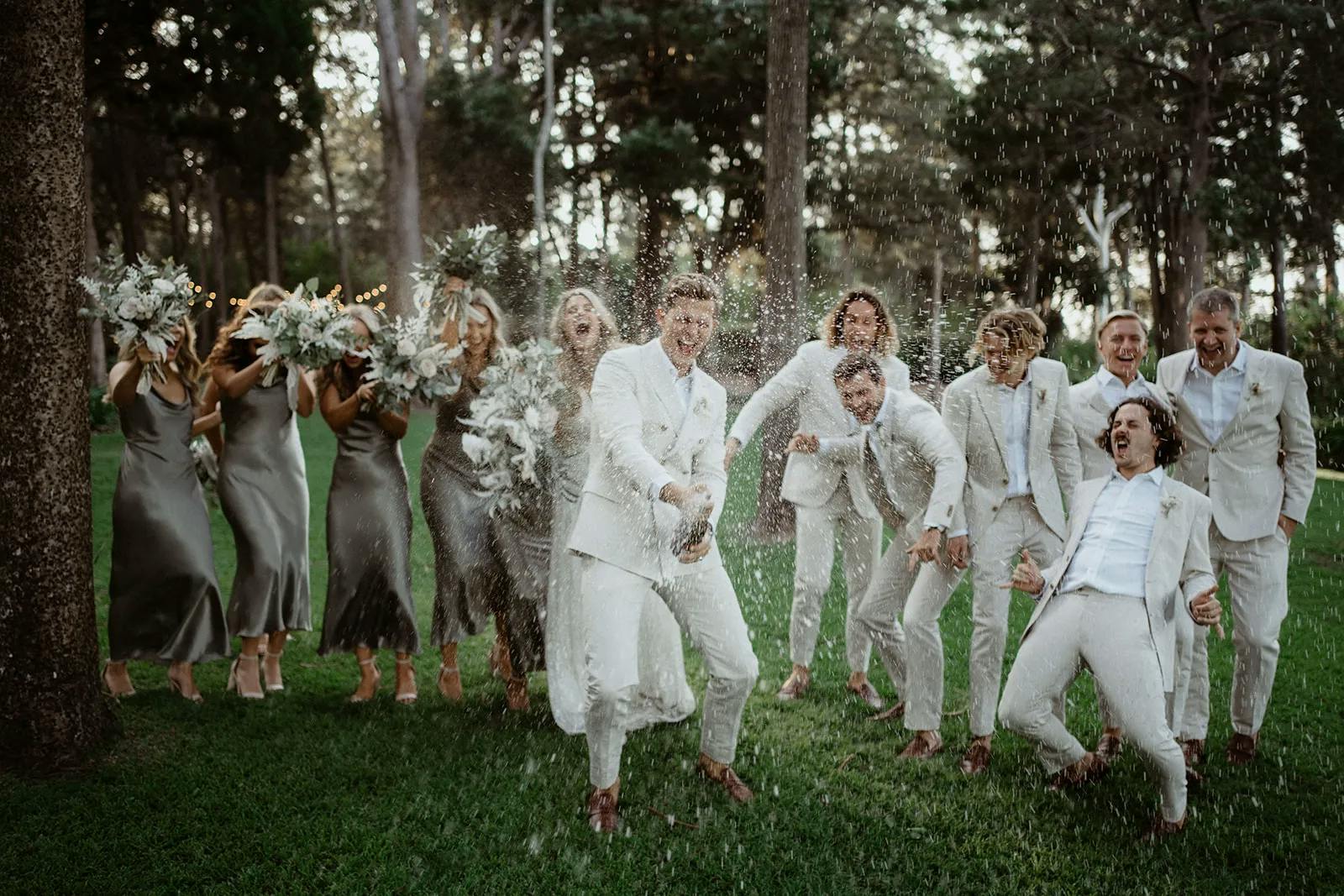 A group of people in formal attire are celebrating outdoors. A person in the center is popping a champagne bottle, causing champagne to spray. Others around them are laughing and reacting joyfully, with some shielding themselves from the spray. Trees are in the background.
