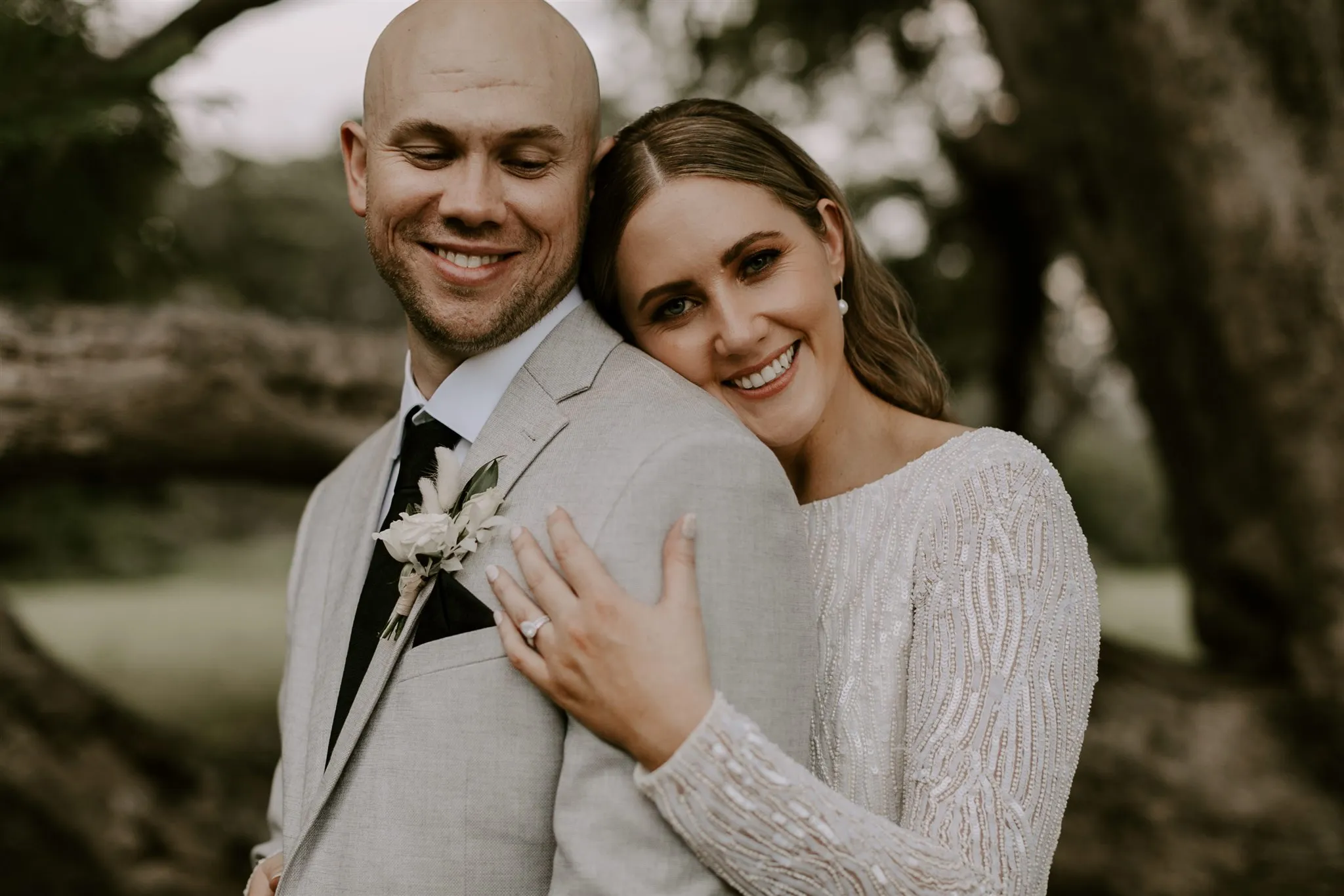 A bride and groom stand closely together outdoors, smiling happily. The bride, in a long-sleeve beaded white dress, has her arms wrapped around the groom from behind. The groom is wearing a light gray suit with a boutonnière and a black tie. Trees are visible in the background.