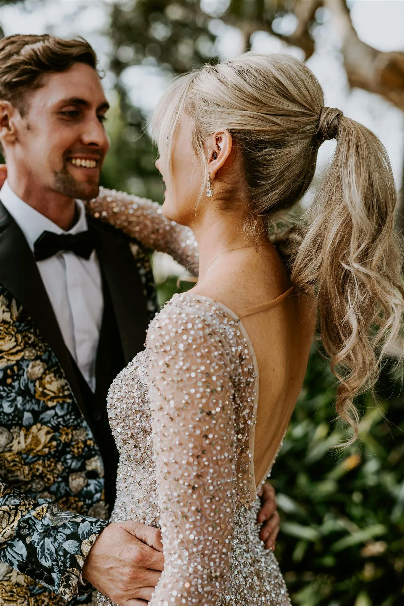 A couple stands close together outdoors, smiling at each other. The woman has long, blonde hair in a ponytail and wears a sparkling, long-sleeved gown. The man has short, brown hair and wears a floral patterned suit jacket with a black lapel and a bow tie.