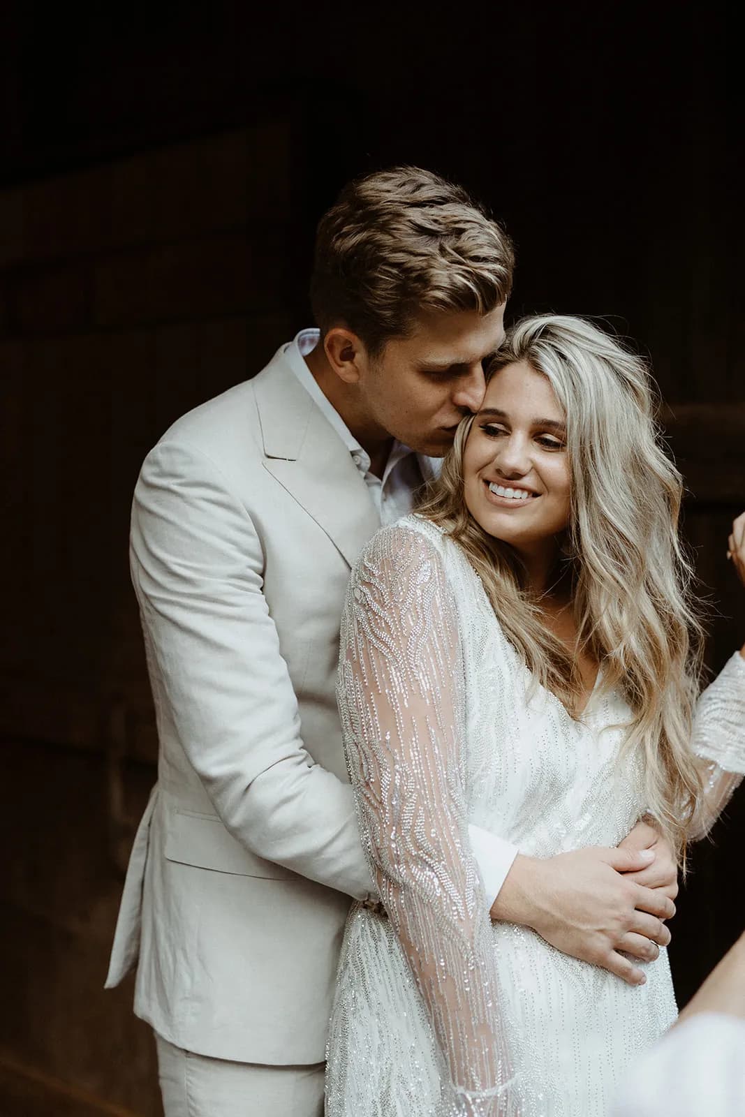A couple dressed in light attire embraces lovingly. The man, in a light suit, stands behind the woman, holding her and gently pressing his face against her head. The woman, in a white dress with sheer sleeves, smiles with her eyes closed.
