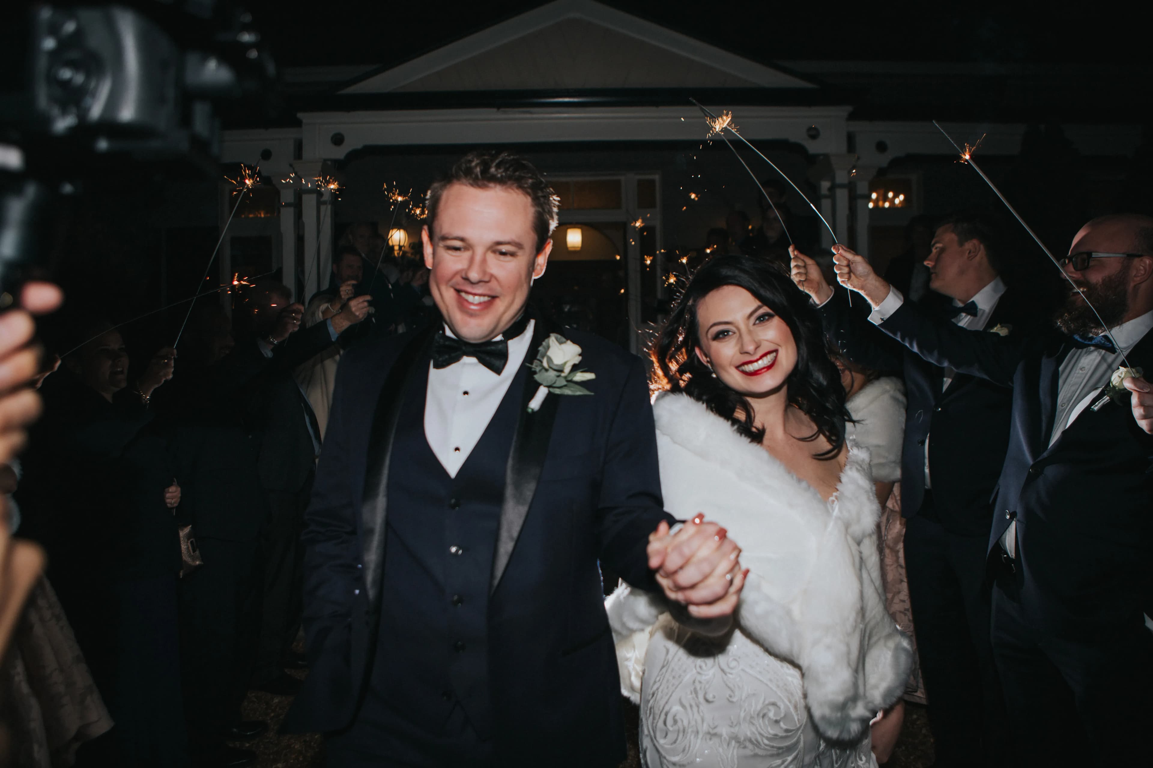 A cheerful couple walks hand-in-hand at night, dressed in formal wedding attire. They smile brightly while surrounded by a crowd holding sparklers, lighting up the celebratory atmosphere. The bride wears a white dress with a shawl, and the groom wears a black tuxedo.