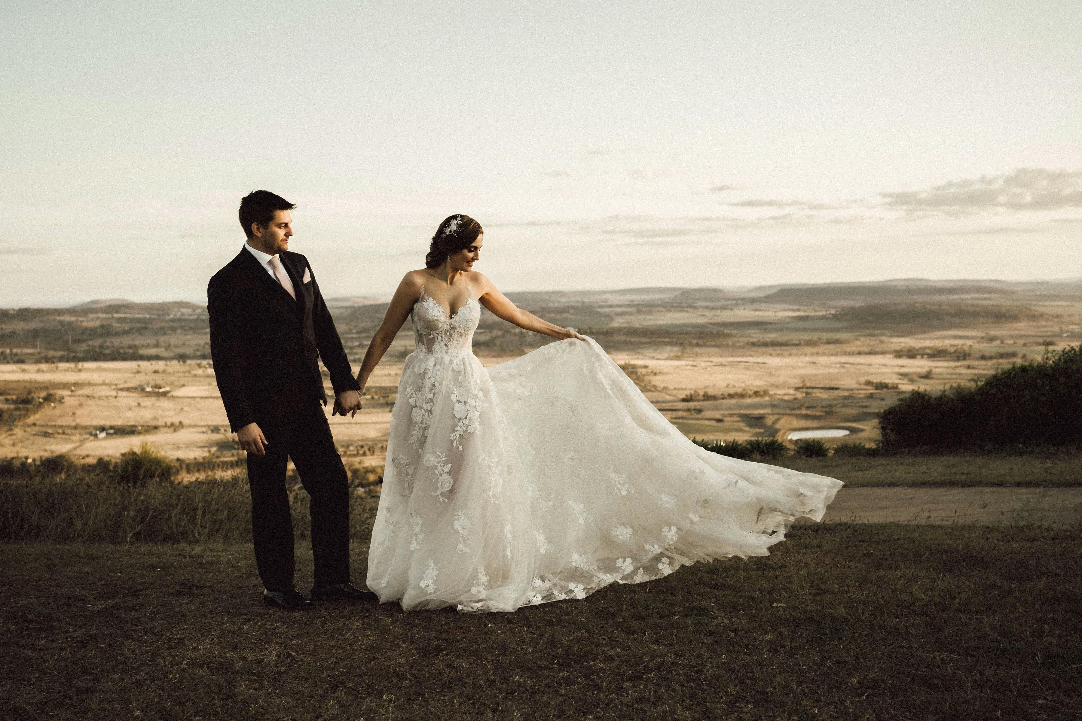A bride and groom stand hand in hand on a grassy hilltop at sunset. The bride is wearing a white strapless gown with lace details and is holding the skirt of her dress. The groom is dressed in a dark suit and tie. The background shows a panoramic view of fields and hills.
