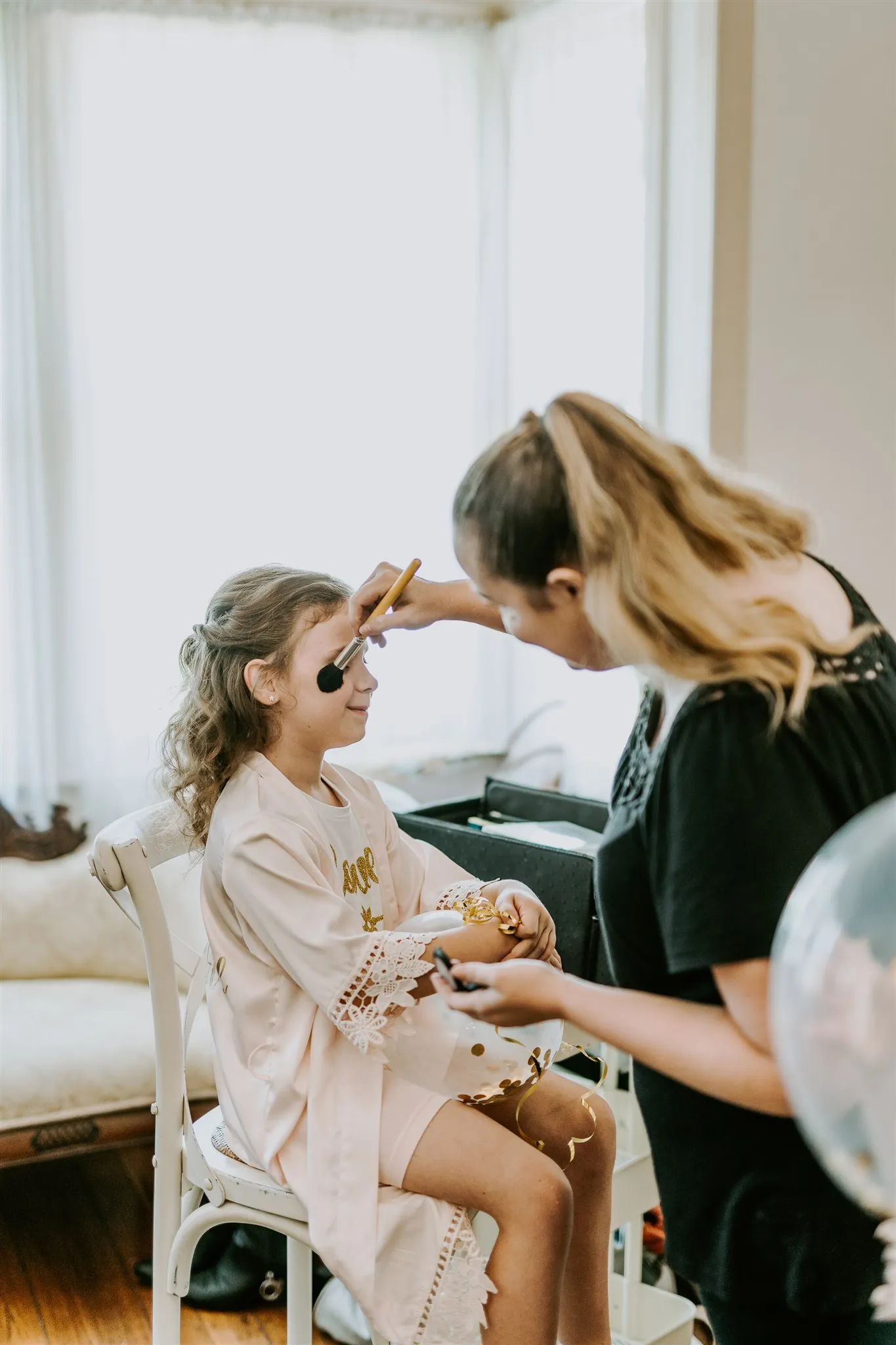 A woman is applying makeup to a young girl who is sitting on a chair. The girl, wearing a pink robe, provides a calm, patient smile as the woman concentrates on her work. The setting appears light and airy, with natural light streaming through large windows.