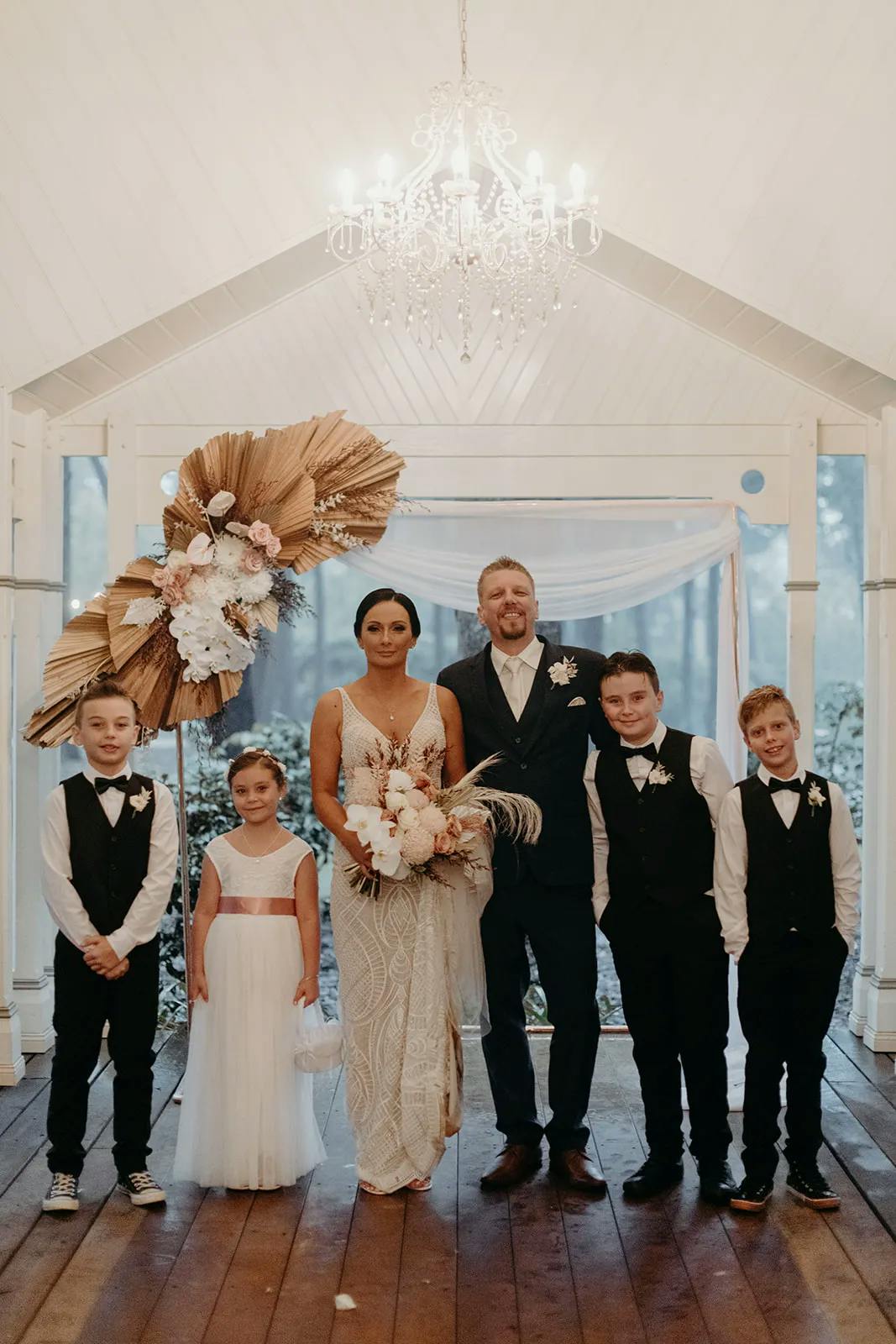 A bride and groom stand together under a chandelier, flanked by four children. The bride holds a bouquet of flowers and wears a white dress, while the groom is in a dark suit with a boutonniere. Two boys stand on each side of the couple, with a girl beside the bride.