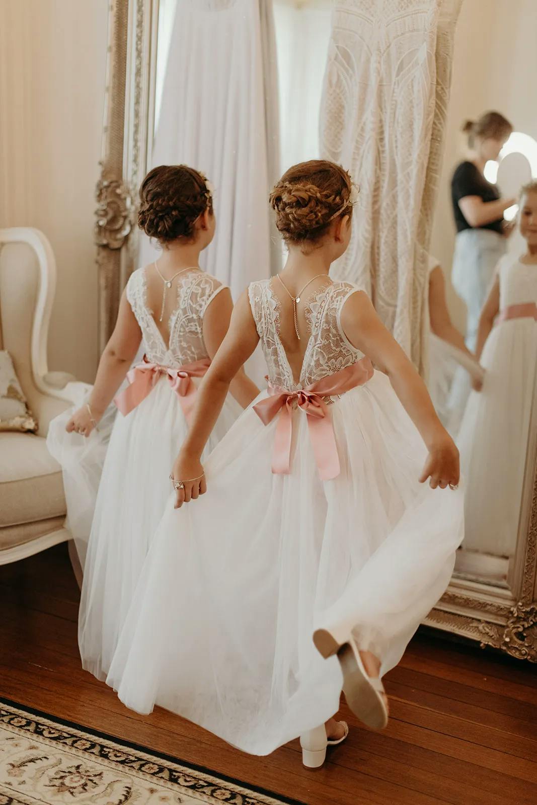 Two young girls wearing white dresses with pink bows stand in front of a large mirror. They have braided hair adorned with floral headbands and are looking at their reflections while gently holding up their dresses. A woman in the background adjusts another girl's dress.