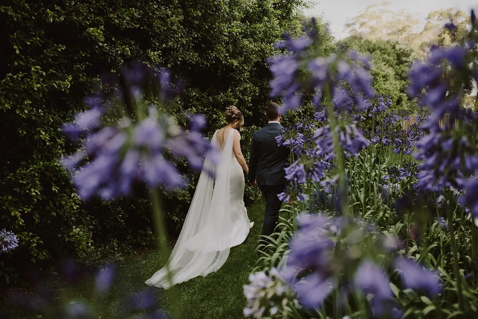 A couple in wedding attire walks hand-in-hand through a garden path lined with tall purple flowers. The bride's flowing veil trails behind her, and the groom is in a dark suit. The scene is lush and green, creating a romantic and tranquil atmosphere.