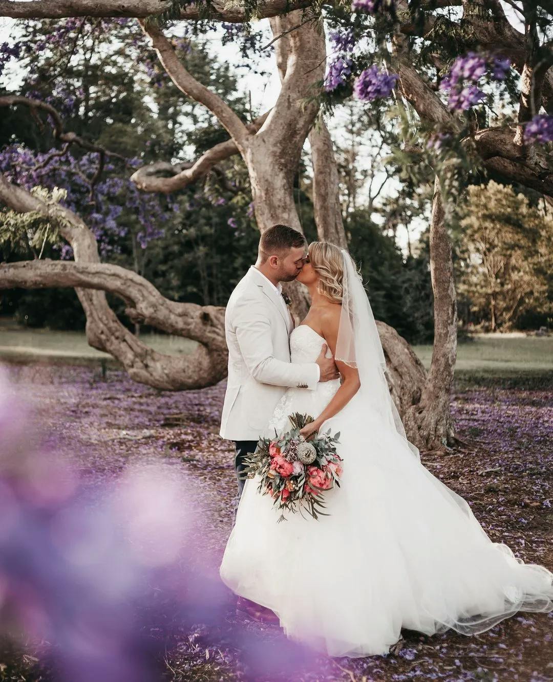 A bride and groom share a kiss under a large tree with twisted branches and purple flowers. The bride holds a bouquet of pink, white, and green flowers and wears a flowing white dress and veil. The ground is covered in purple petals, creating a whimsical atmosphere.