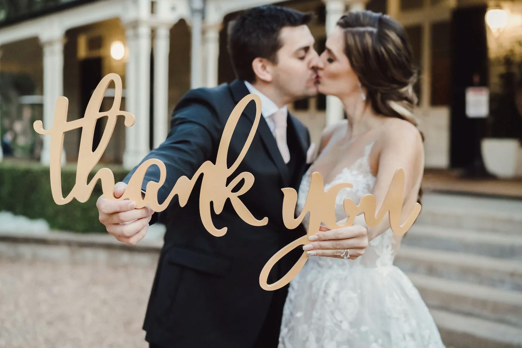 A bride and groom, in wedding attire, share a kiss while holding a decorative sign that says "thank you" in cursive letters. The background features a building with an elegant outdoor setting.