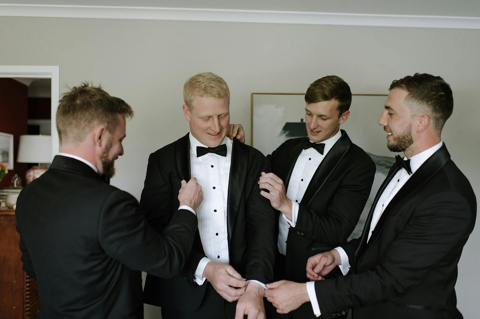 Four groomsmen in tuxedos are adjusting the groom's attire and bow tie. They are inside a room with light-colored walls and a piece of framed art in the background. The atmosphere appears joyful as they prepare for a formal event.