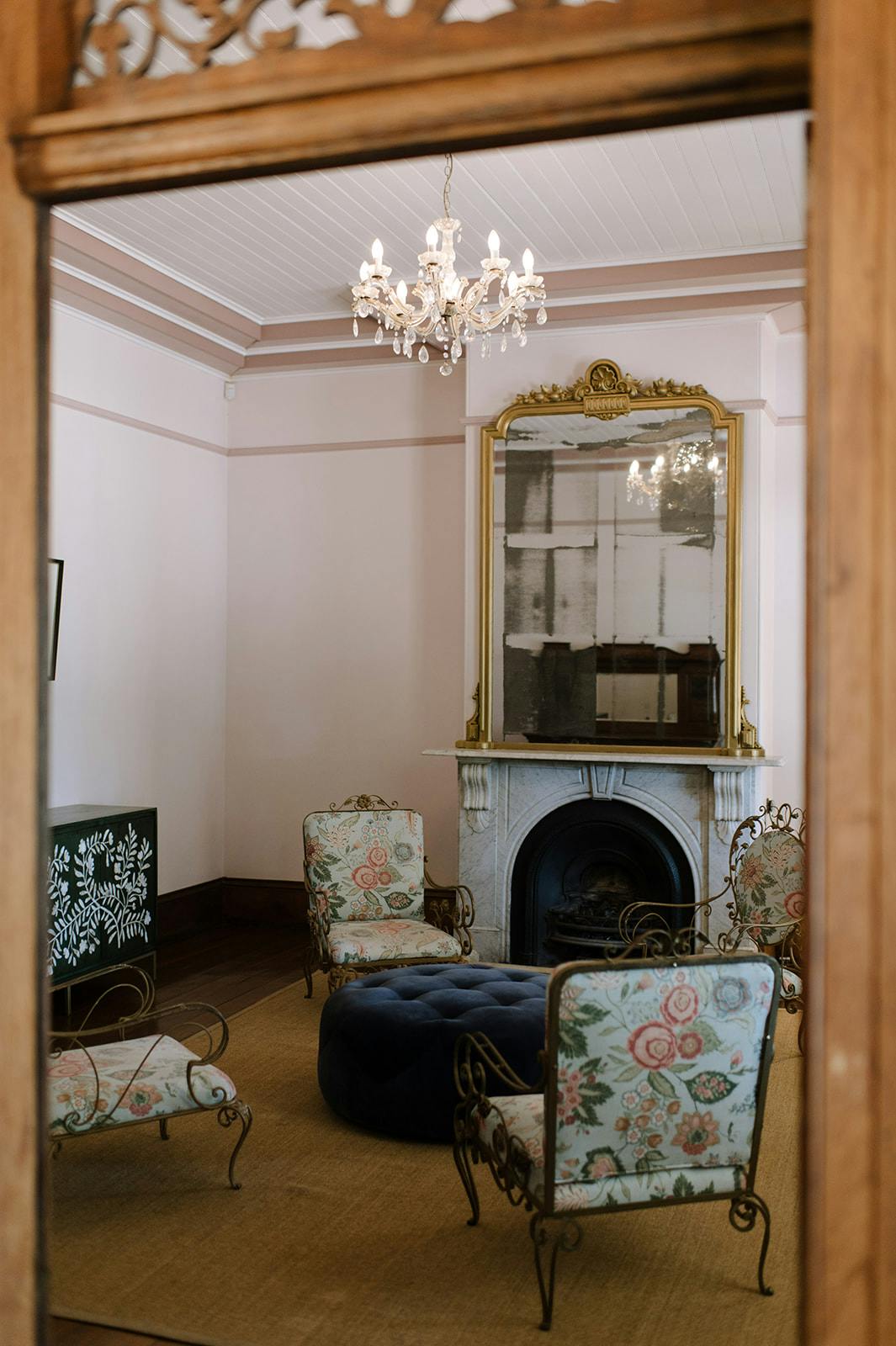 A cozy living room with ornate furniture, including four floral-upholstered chairs surrounding a large blue ottoman. The room features a fireplace with a tall mirror above it and a chandelier hanging from the ceiling. The decor is elegant with a vintage touch.