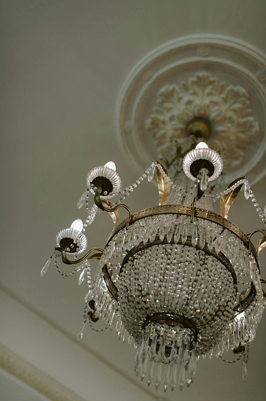 A close-up view of an ornate crystal chandelier with multiple tiers of hanging crystals, intricately designed metal arms, and candle-shaped light bulbs. The ceiling above features decorative molding. The chandelier is illuminated, casting a soft light.