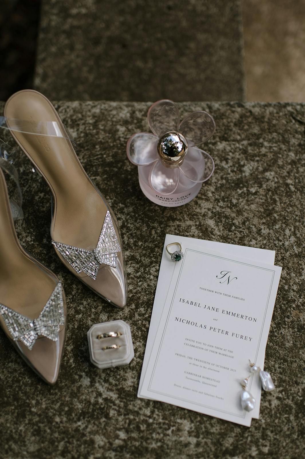 A flat lay photo of wedding items: a pair of transparent and jeweled pointed-toe high-heeled shoes, a bottle of Marc Jacobs Daisy perfume, a wedding invitation, pearl earrings, and two wedding rings in a box. The setting is an outdoor stone surface.