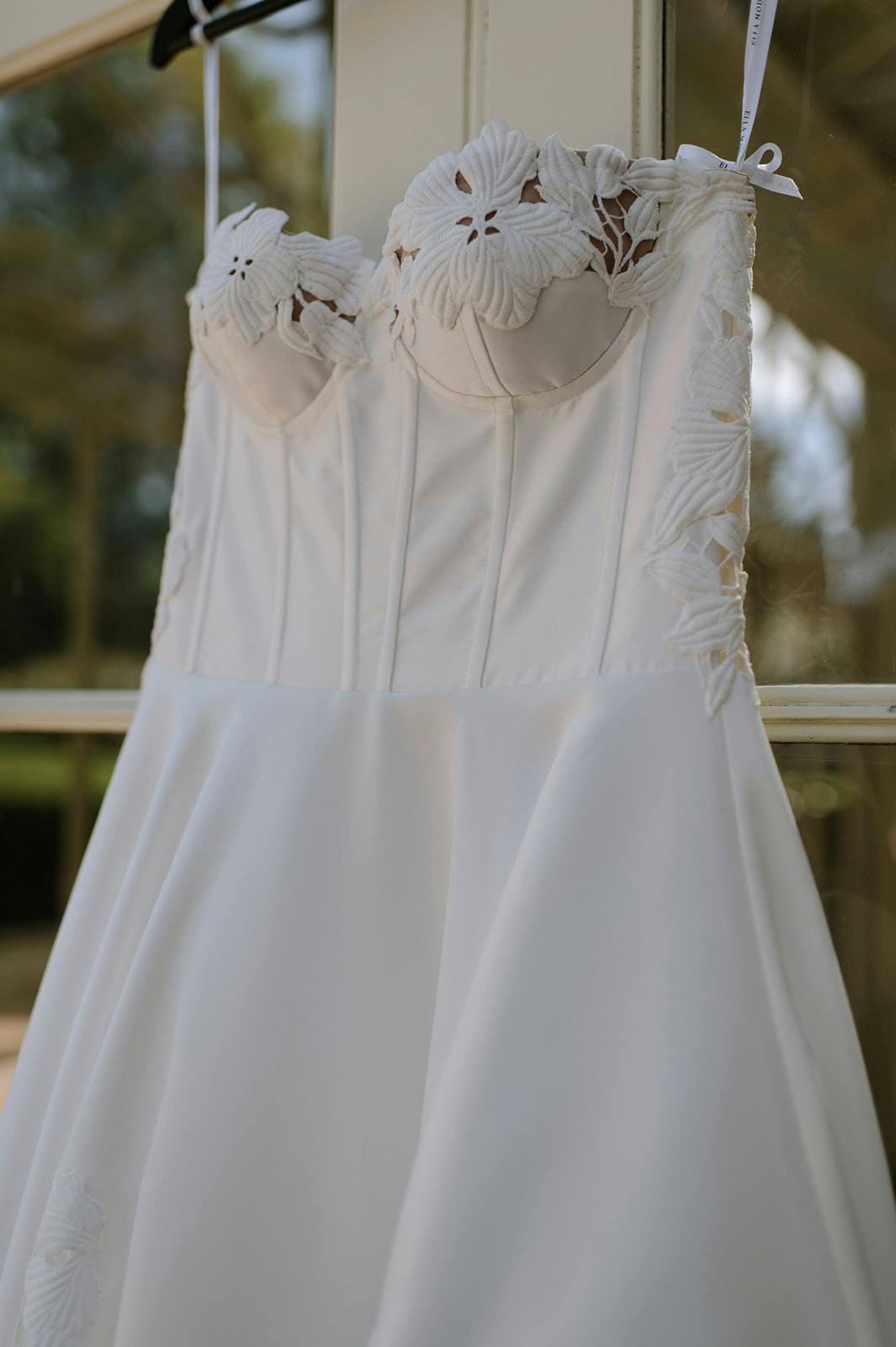 A close-up of a white strapless wedding dress hanging on a hanger. The dress has floral lace detailing around the bust and on the edges of the bodice. The fabric is smooth and the overall design of the dress is elegant and sophisticated.