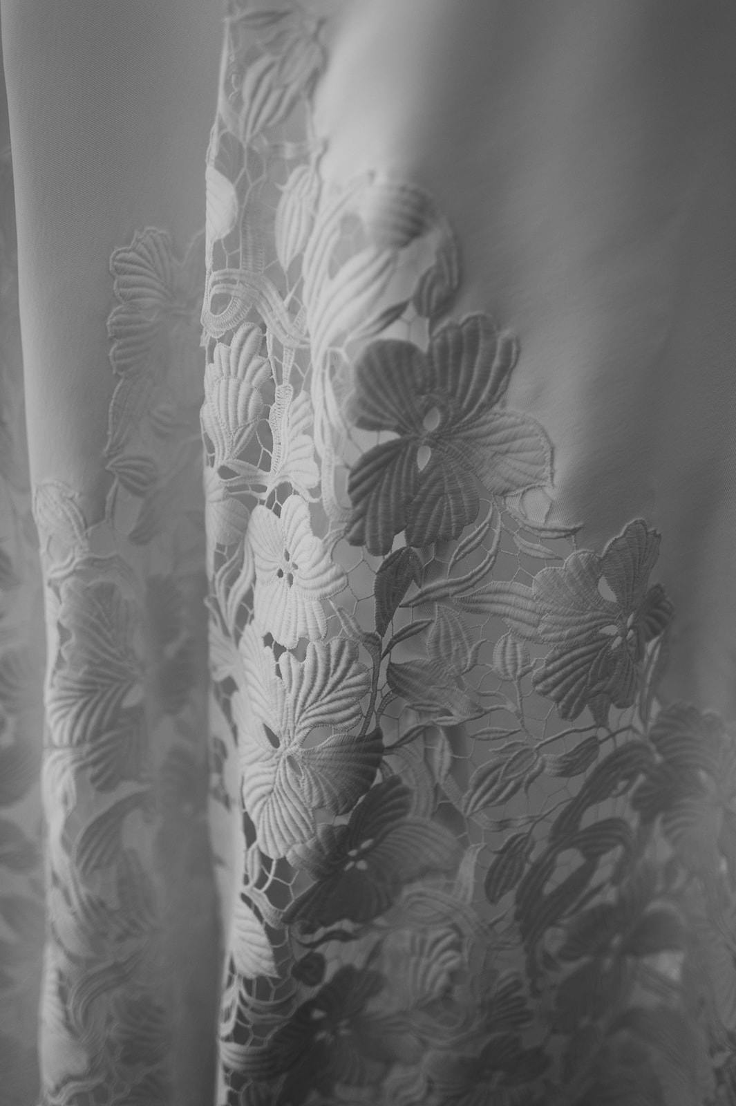 Black and white close-up of a delicate lace fabric featuring intricate floral patterns. The lace appears to be draped, creating soft, flowing folds with varying light and shadow effects.