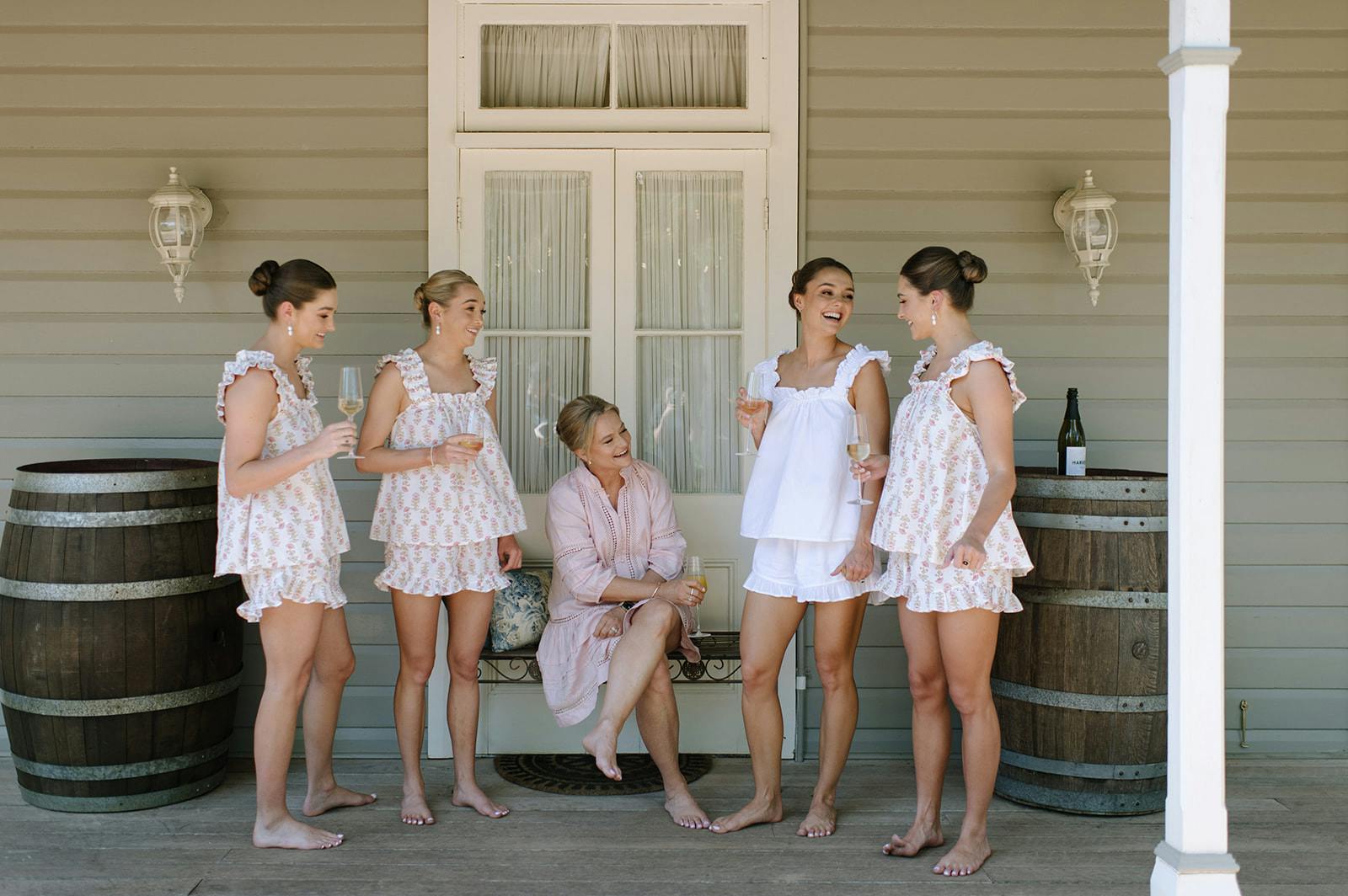 A group of five women, dressed in matching floral pajamas, gather on a wooden porch. One woman sits on a chair holding a drink, while the others stand and laugh, holding glasses. They appear to be enjoying a casual and joyful moment together.