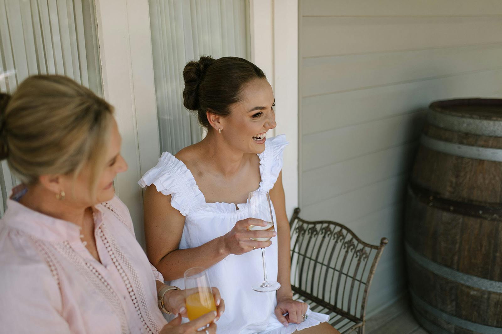 Two women enjoying drinks on a porch. The woman on the right, in a white sleeveless dress, holds a wine glass and laughs. The woman on the left, wearing a light pink blouse, holds an orange drink. They sit near a wooden barrel and a metal chair.
