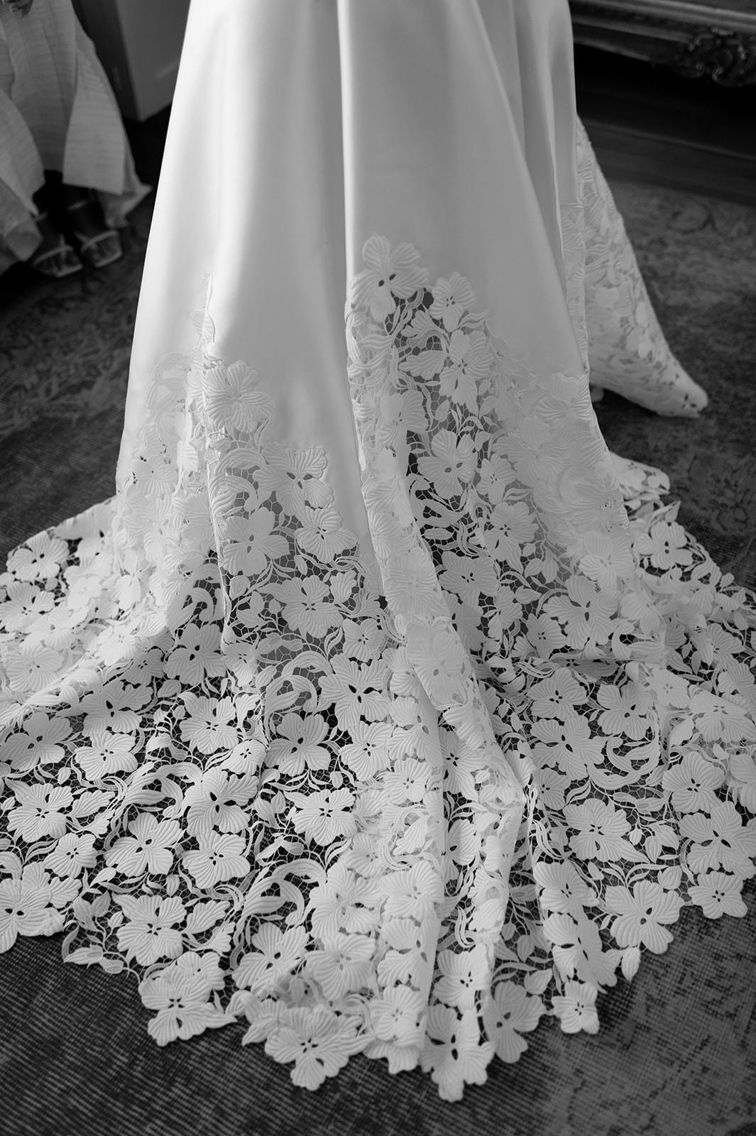 A close-up black and white image of the lower part of a wedding dress featuring intricate lace details with floral patterns extending from the hem. The dress is elegantly pooled on the floor, highlighting the delicate craftsmanship of the lacework.