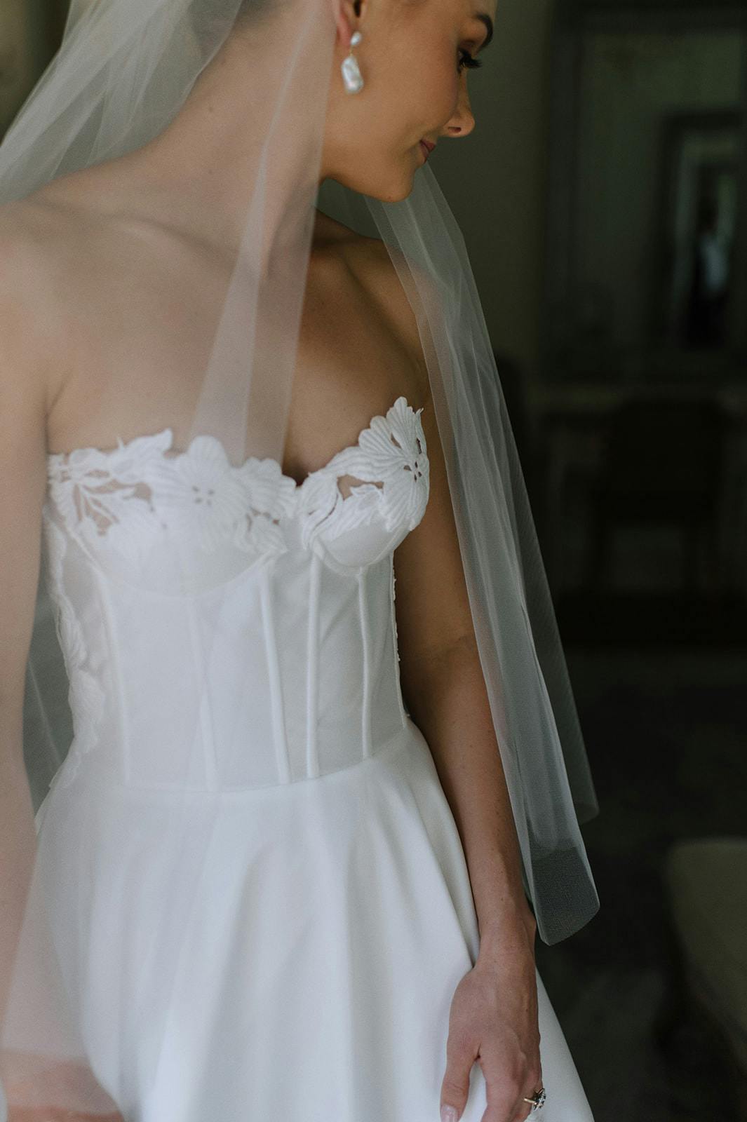 A bride stands in a white corset-style wedding dress adorned with lace flowers, wearing a delicate veil and pearl earrings. She has a hand on her hip, and her head is turned to the side, looking down with a smile. The background is softly blurred.
