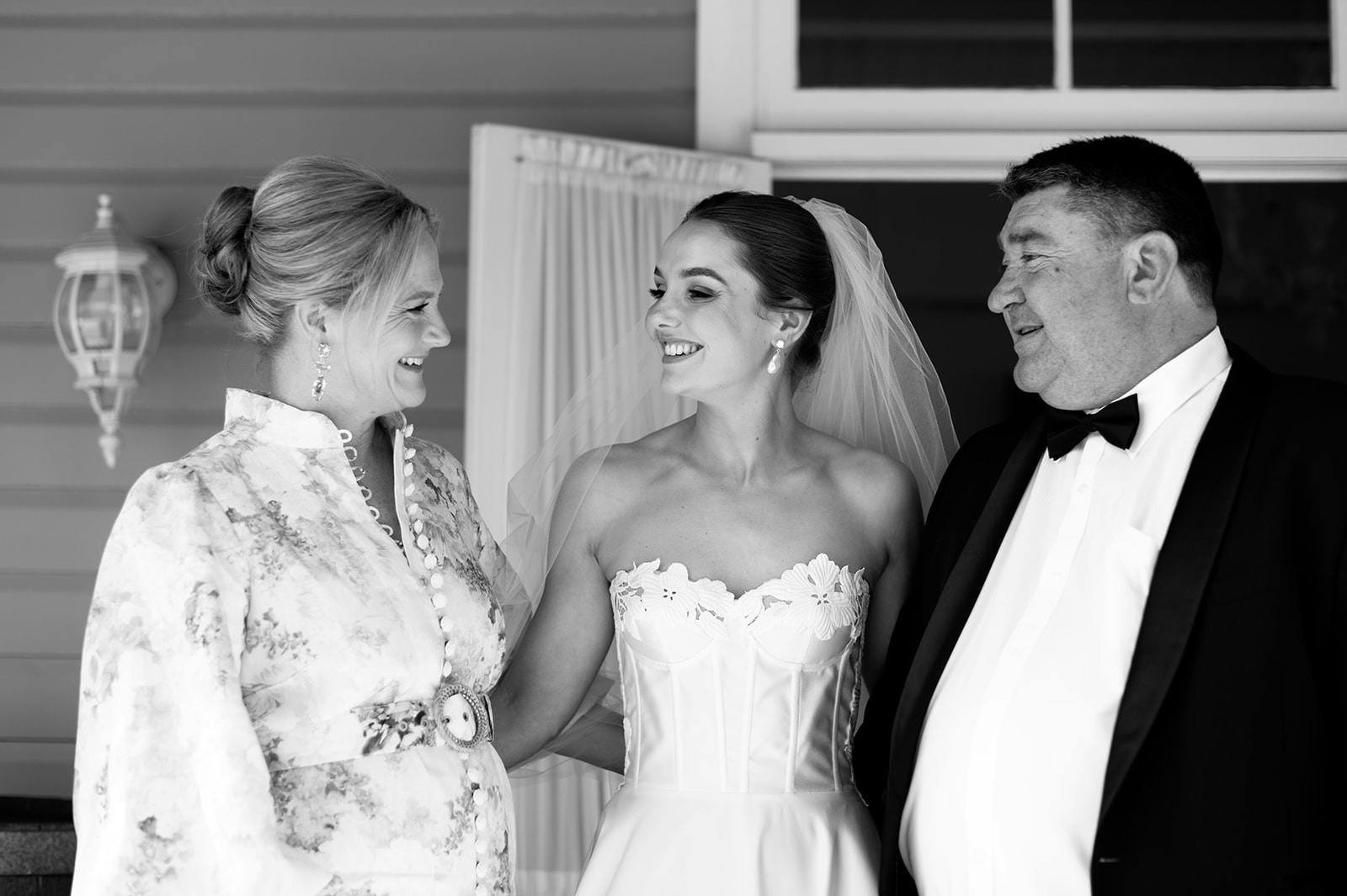 A bride in a strapless wedding dress and veil smiles, looking at a woman in a floral dress and a man in a tuxedo. They both smile back at her. They appear to be standing on a porch with a window in the background.