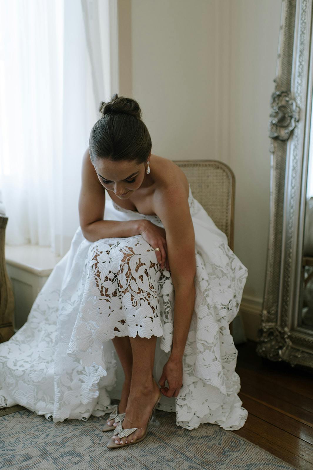 A bride in a white lace wedding dress sits on a wicker chair, bending down to adjust her shoe. She has her hair styled in an elegant updo and is in a softly lit room with a large window featuring sheer white curtains. An ornate large mirror stands next to her.