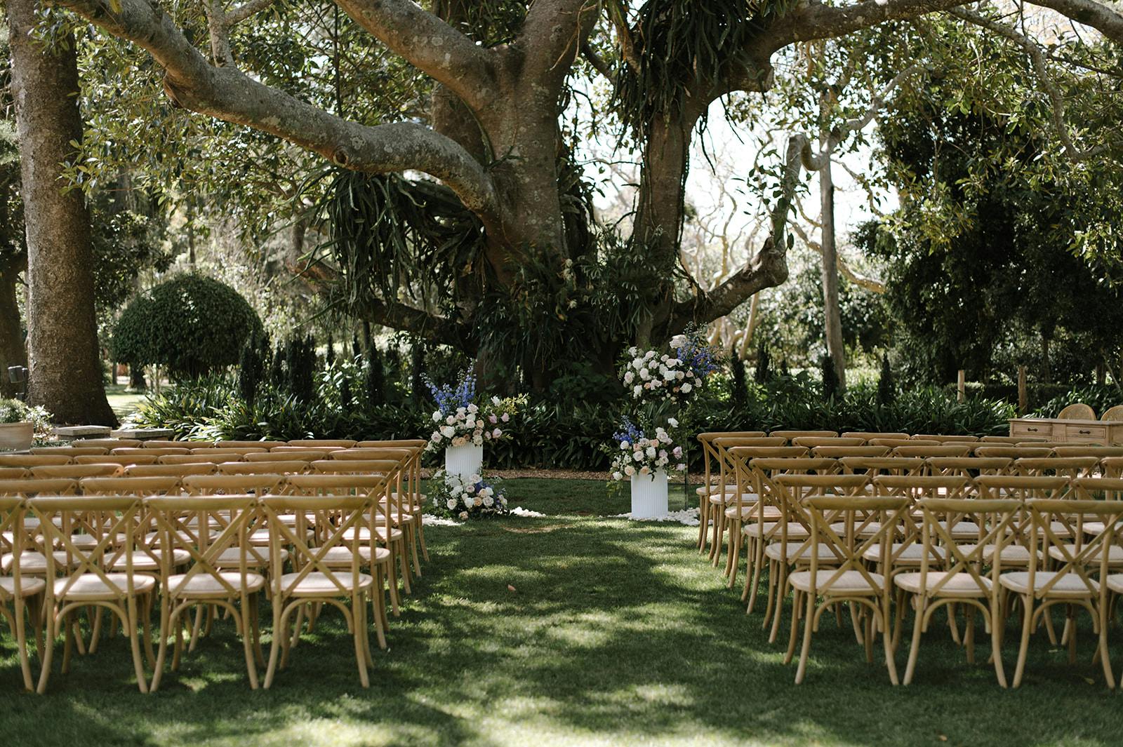 An outdoor wedding setup featuring wooden chairs arranged in rows on green grass, leading to a large tree. The tree is adorned with floral arrangements in shades of white, pink, and purple, creating a natural archway. The scene is surrounded by lush greenery.