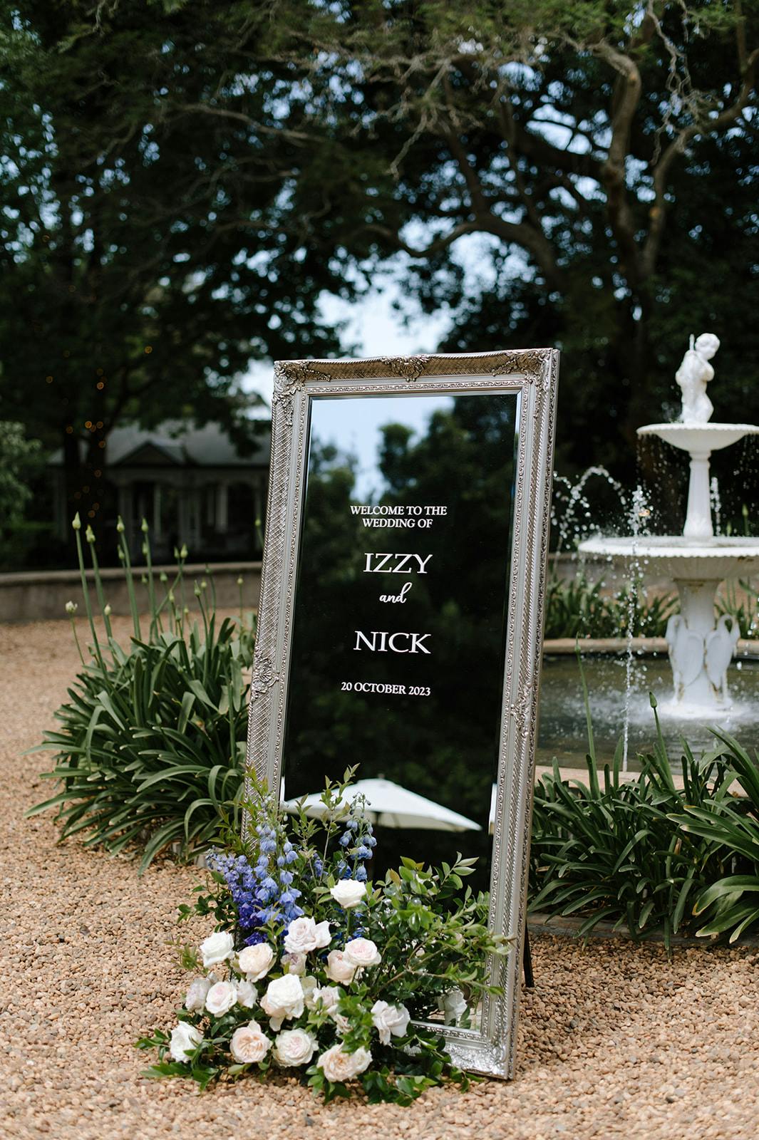A decorative mirror with a silver frame stands outdoors, displaying white text that reads: "Welcome to the Wedding of Izzy and Nick, 20 October 2023." The mirror is adorned with a floral arrangement of white and purple flowers at its base, near a fountain.