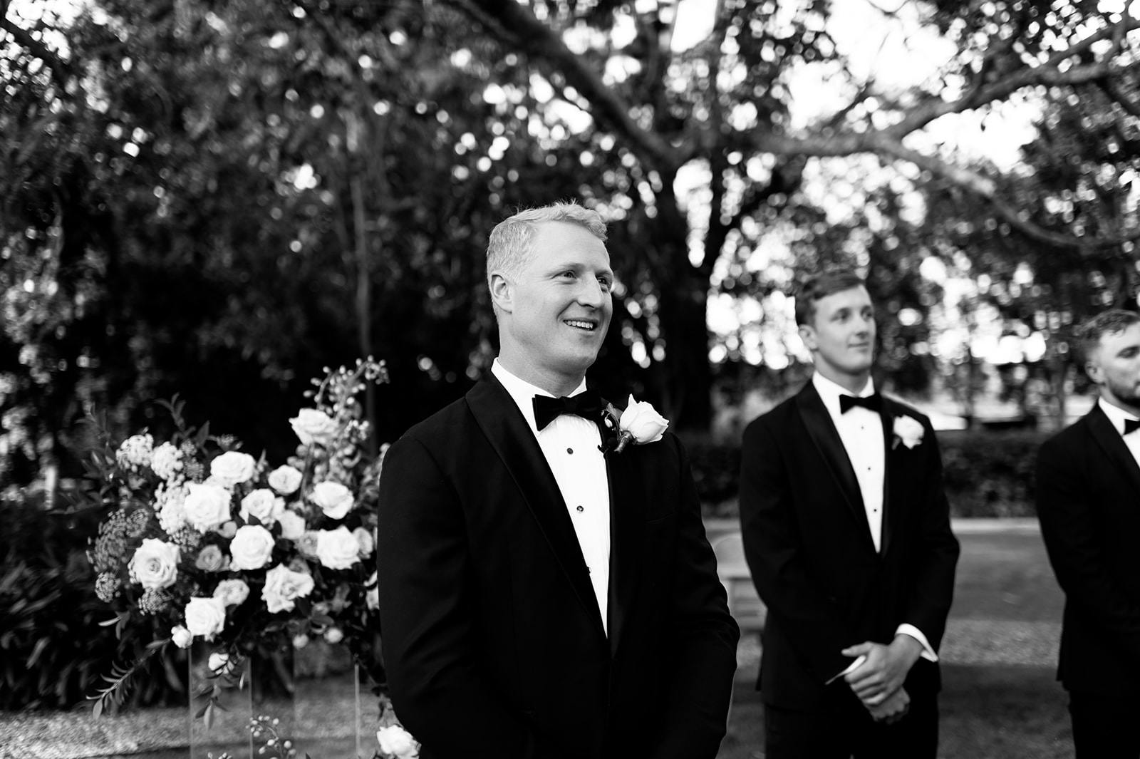 A groom in a tuxedo smiles happily while standing outdoors next to two groomsmen, also in tuxedos. Behind them, there is a lush garden area with trees and flowers. The scene is festive, and the men look poised and joyful.