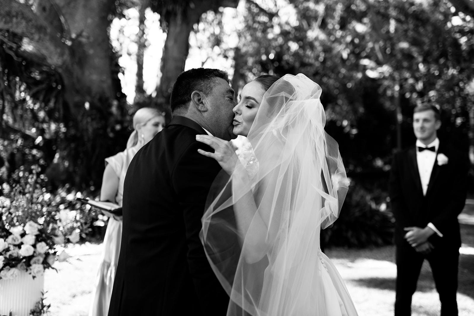 A bride and groom share an emotional embrace and kiss during their outdoor wedding ceremony. The bride is wearing a veil and the groom is dressed in a suit. Bridesmaids and groomsmen stand in the background surrounded by greenery and floral arrangements.