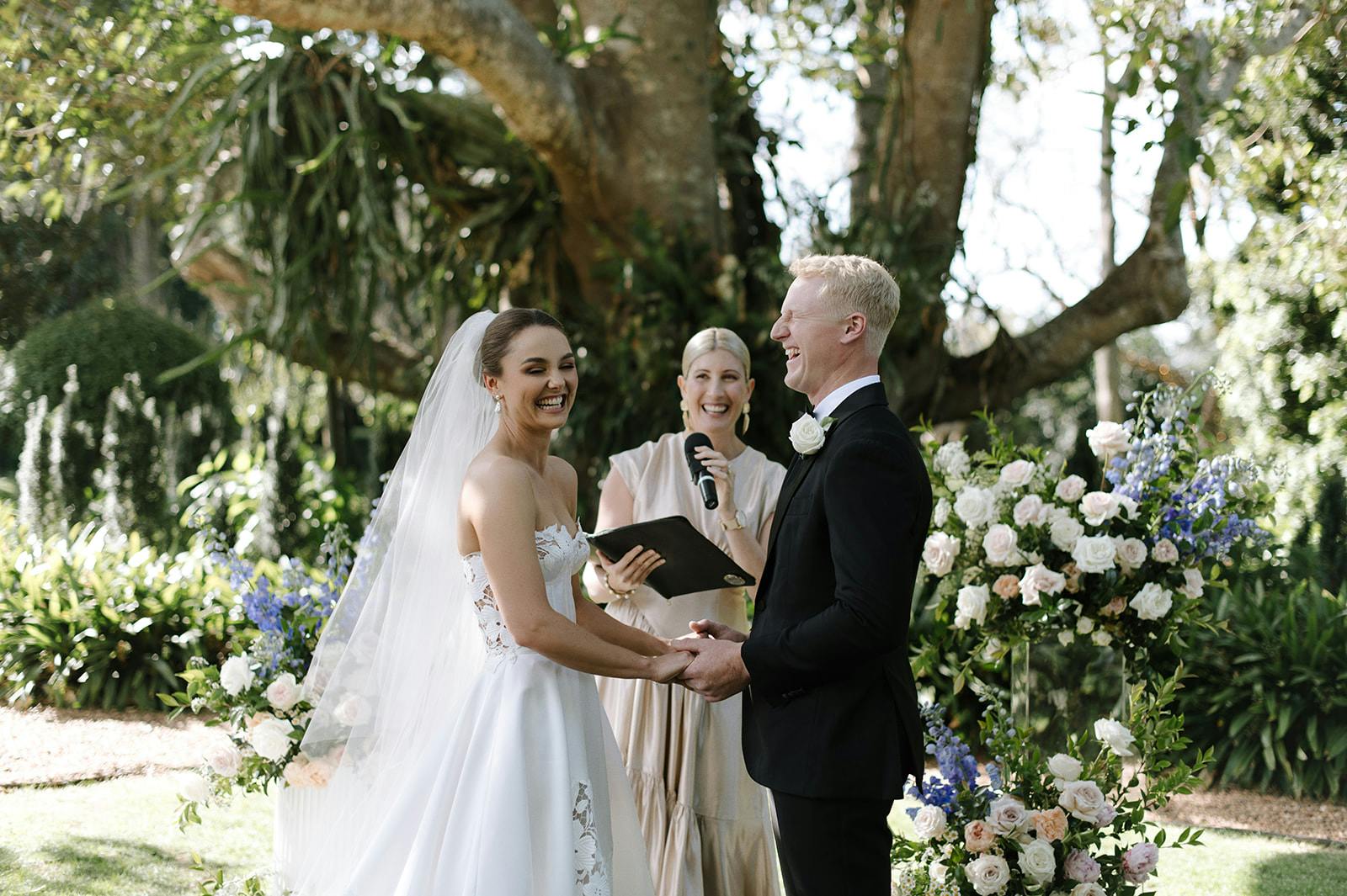 A bride and groom, holding hands and smiling at each other, stand facing an officiant during their wedding ceremony outdoors. The bride is in a white gown with a veil, and the groom is in a black suit. The background is lush with greenery and floral arrangements.