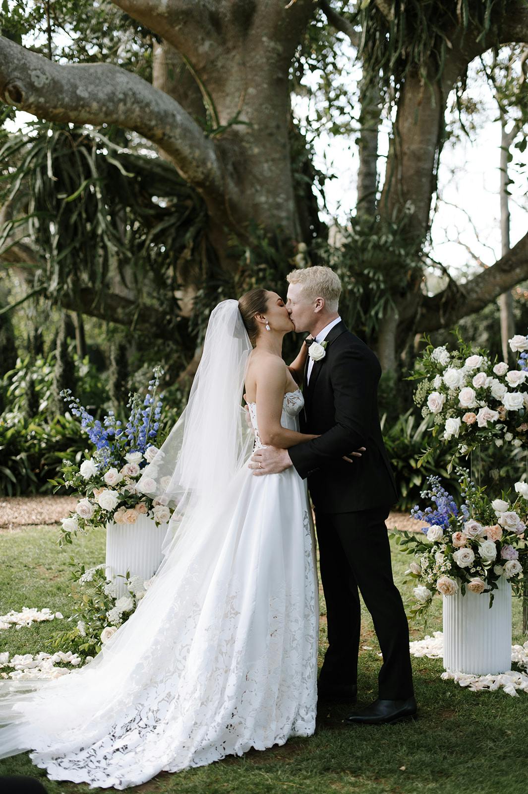 A bride and groom share a kiss in an outdoor wedding ceremony, standing in front of a large tree adorned with greenery. The bride is wearing a white, lace wedding gown with a veil, and the groom is in a black suit with a white boutonniere. Floral arrangements surround them.