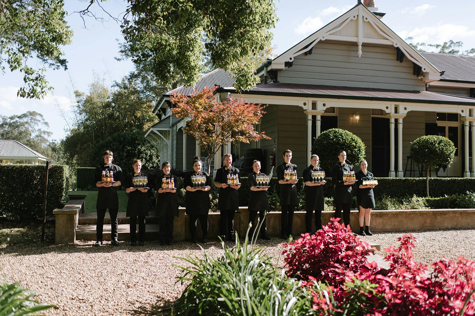 A group of people in matching black attire stands in a row outdoors, each holding a glass of champagne. They are in front of a charming, grey wooden house with white trim, surrounded by well-maintained garden plants and trees. The scene is sunny with a clear blue sky.