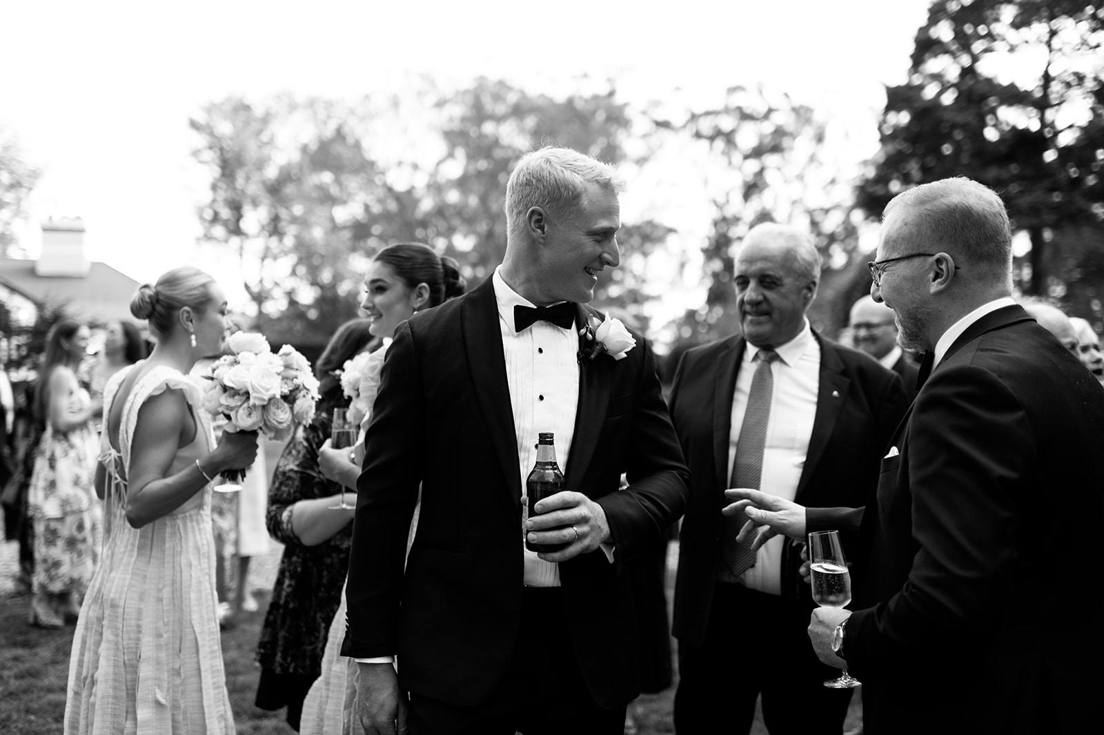 Black and white photo of a group at an outdoor event. A man in a tuxedo holds a bottle, smiling at a man in a suit holding a glass. Behind them, women chat and hold flowers. Trees and other guests fill the background.
