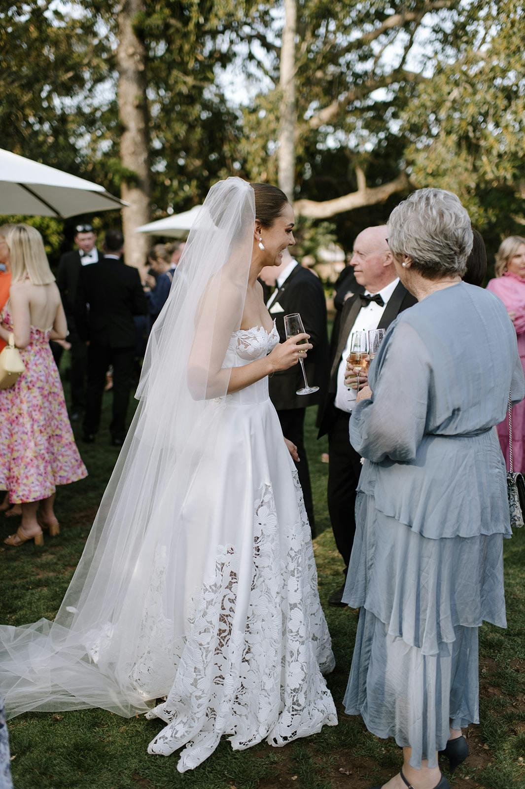 A bride in a white wedding gown with lace details and a long veil converses with two older guests, one in a light blue dress, holding champagne glasses. Other guests in formal attire and umbrellas are visible in the background at this outdoor event.