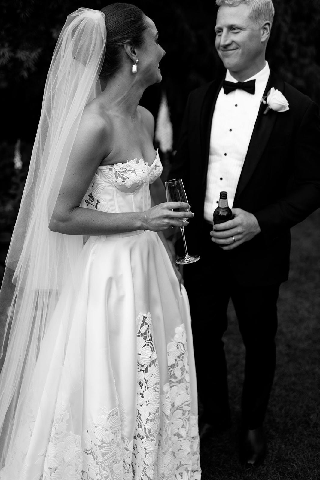 A bride and groom share a joyful moment. The bride, in a strapless wedding dress with a veil, holds a glass of wine. The groom, wearing a tuxedo and bow tie, holds a bottle of beer. They are smiling at each other against a dark, outdoor background.