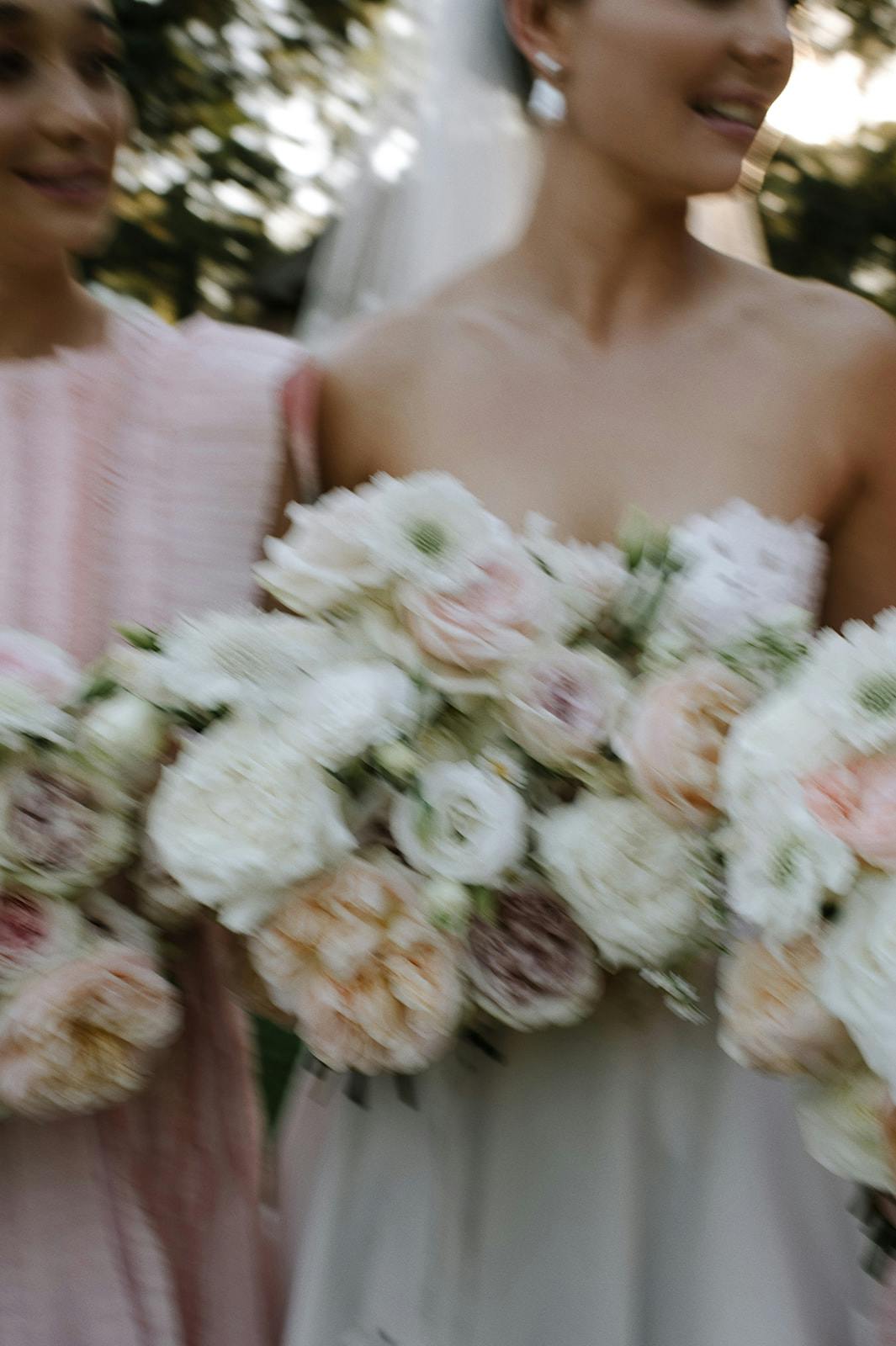 A blurred image shows two people standing next to each other, holding large bouquets of white and pastel-colored flowers. The person on the right is wearing a white strapless dress, while the person on the left is in a light pink outfit.