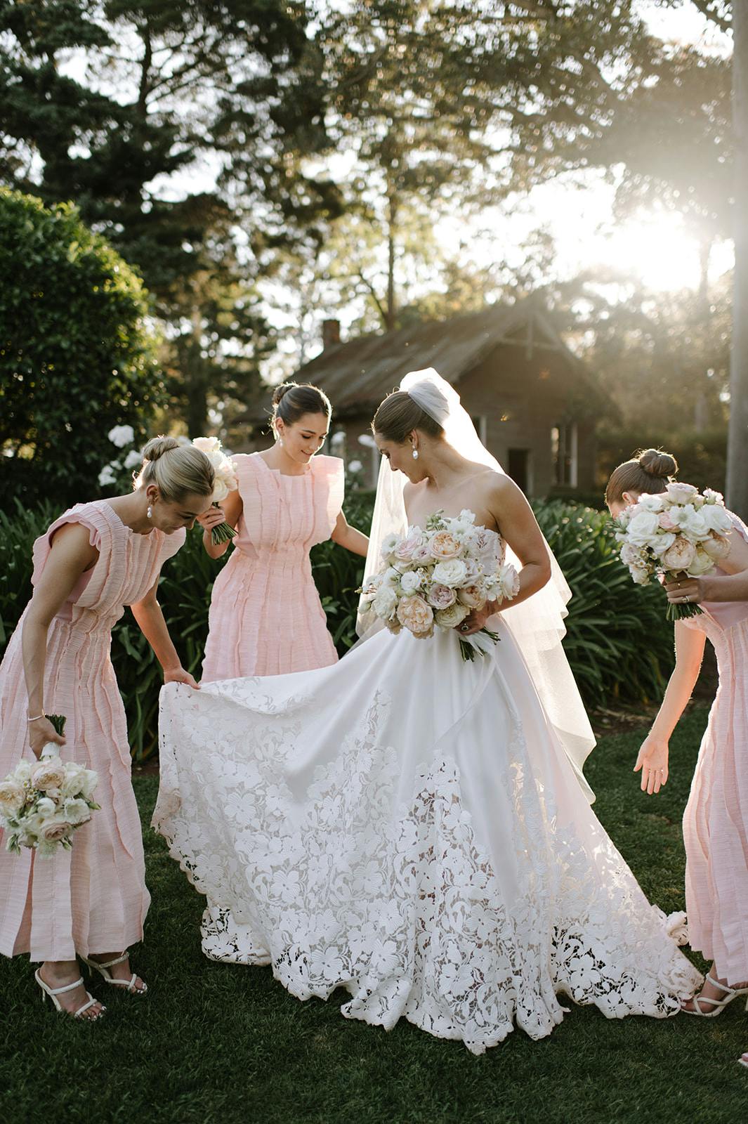 A bride in a white lace wedding gown and veil is surrounded by three bridesmaids in light pink dresses. They are outdoors on a grassy area with sunlight filtering through trees. The bridesmaids are helping to arrange the bride's dress as she holds a bouquet of white flowers.