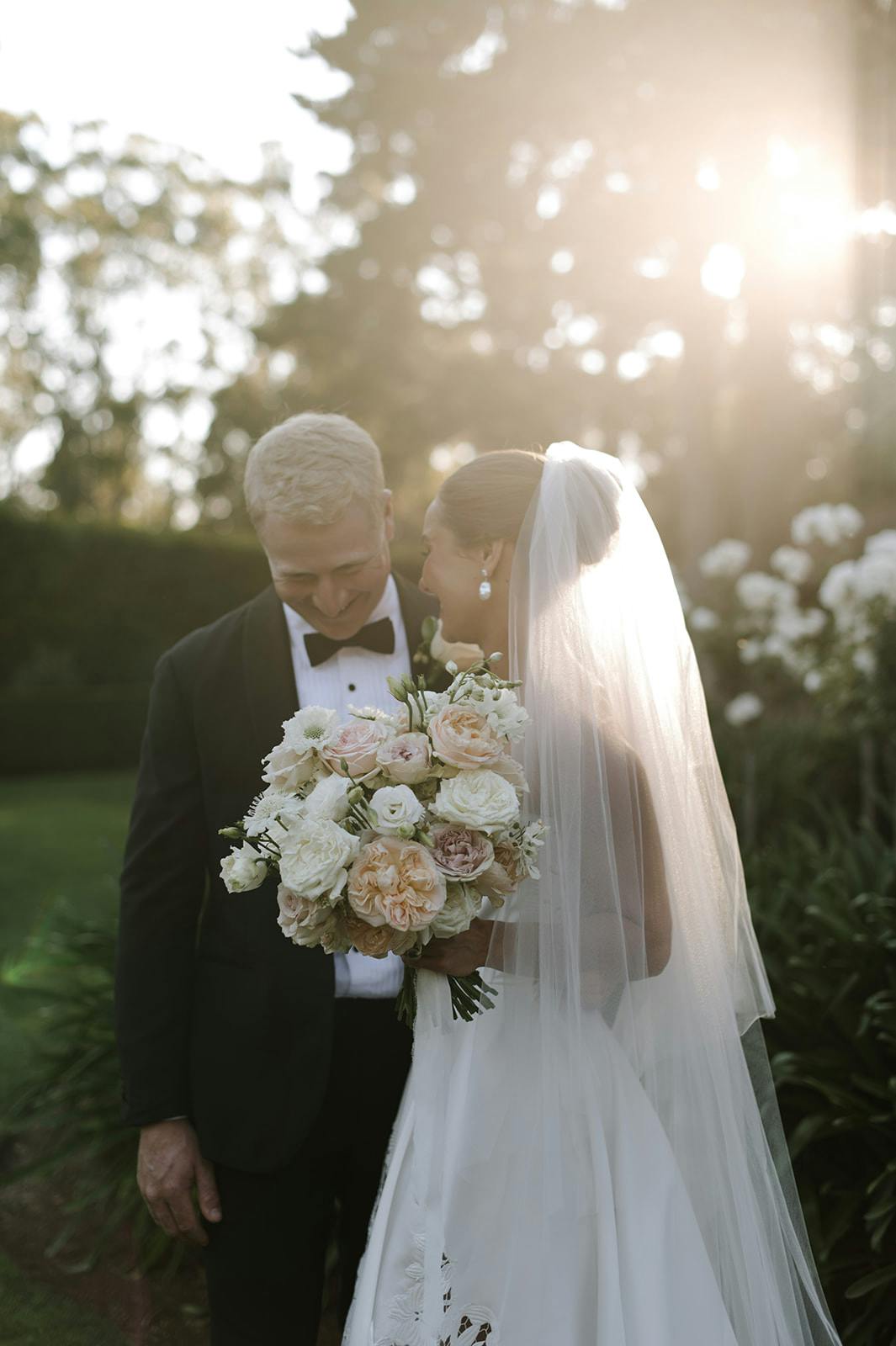 A bride and groom share a tender moment outdoors during their wedding. The bride holds a bouquet of white and peach flowers and wears a veil, while the groom looks down, smiling. Sunlight filters through the trees, creating a warm, golden glow around them.