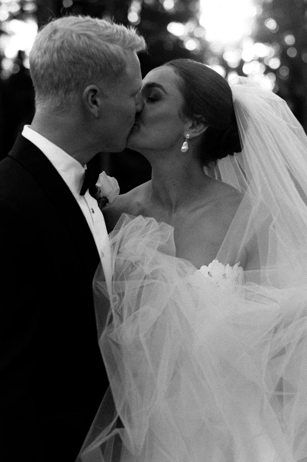 A bride and groom share a kiss outdoors. The bride is wearing a strapless gown with tulle and a veil, accompanied by drop earrings. The groom is in a tuxedo with a boutonnière. The black-and-white image has blurred background foliage and bright light filtering through.
