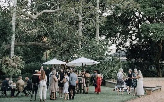 A group of people mingles outdoors in a lush garden setting. Some are seated at tables, while others stand and chat near white umbrellas. Tall trees and greenery surround the gathering, creating a serene and natural atmosphere.