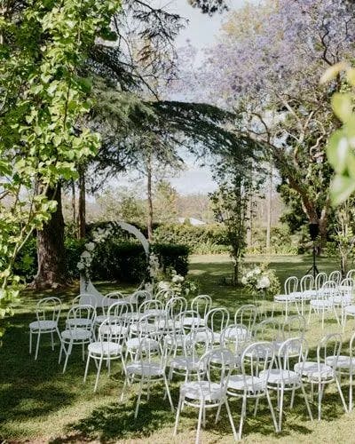 A serene outdoor wedding setup in a lush garden with rows of white chairs arranged before a white floral arch. The garden is surrounded by tall trees, sunlight filters through the leaves, and the atmosphere is calm and inviting.