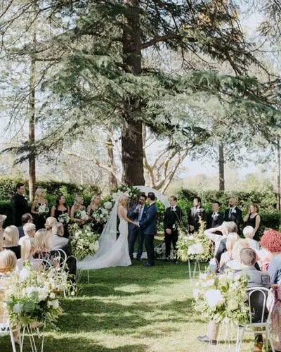 A wedding ceremony is taking place outdoors under a large tree. The bride and groom stand facing each other, surrounded by their wedding party. Guests are seated in rows on the grass, observing the ceremony. Greenery and floral arrangements decorate the scene.