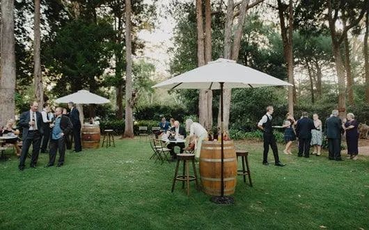 A garden party with people standing and sitting under white umbrellas at tables. The setup includes barrel tables and surrounded by trees and greenery. Some guests are mingling and chatting, while others are seated, all dressed in formal attire.