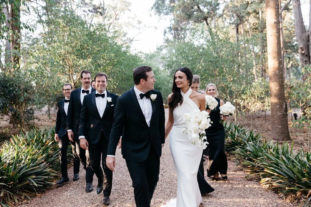 A bride and groom walk down a path outdoors, dressed elegantly in a white gown and black suit, respectively. They are accompanied by a smiling bridal party in formal black attire. The scene is set in a lush, green garden with trees and foliage in the background.