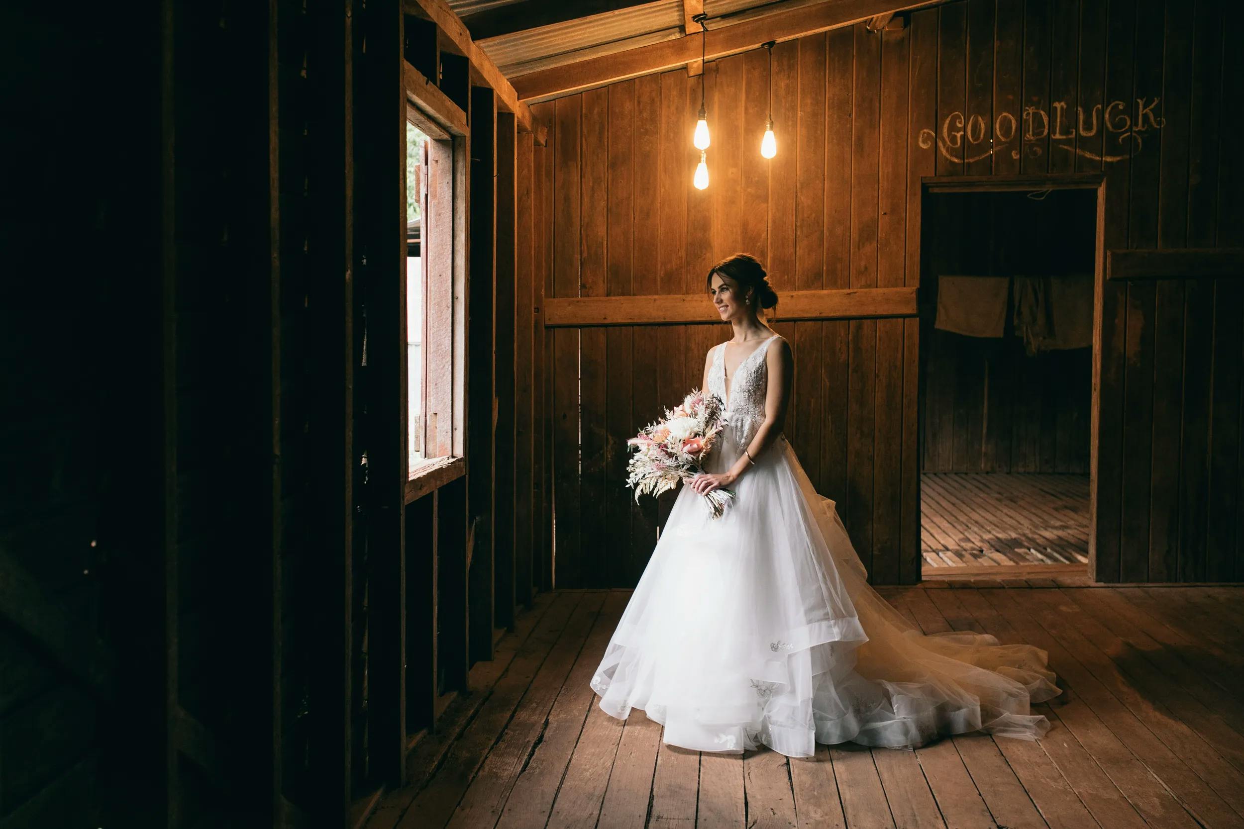 A bride in a white wedding dress stands inside a rustic wooden room with exposed beams, holding a bouquet of flowers. She gazes out of a window on her left as warm light bulbs hang from the ceiling, casting a soft glow. A sign reading "Good Luck" is on the back wall.