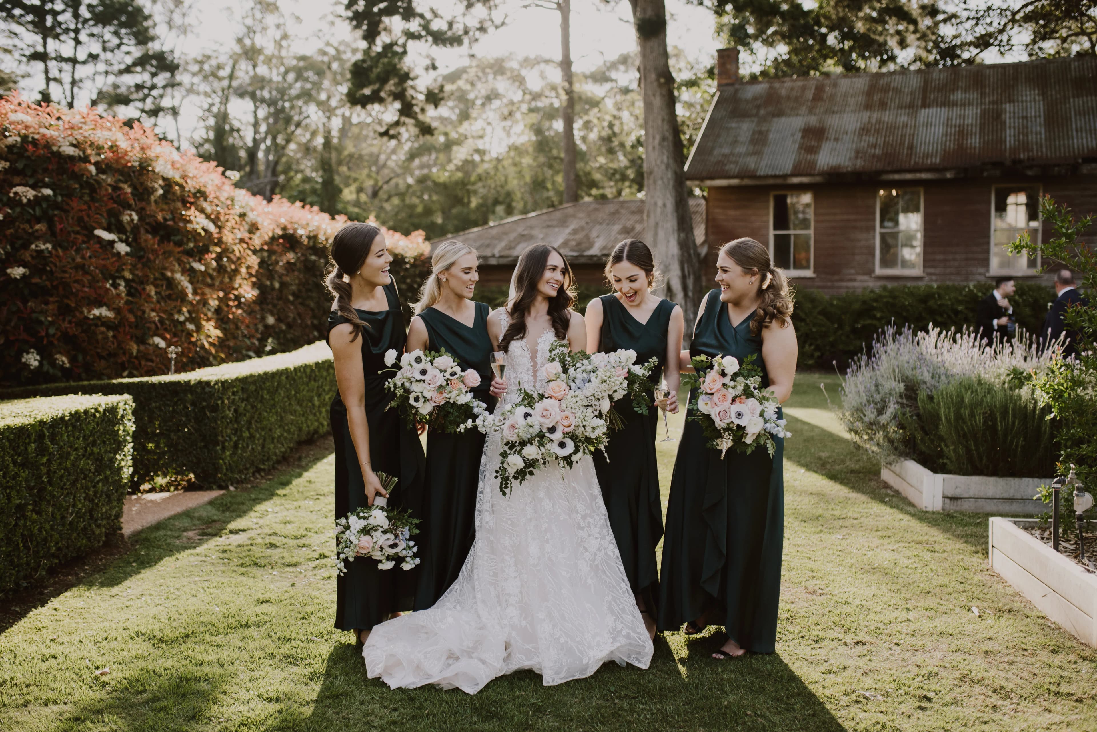 Five women stand together in a garden, smiling and holding bouquets of flowers. The woman in the center wears a white dress, while the other four wear dark green dresses. A rustic house and trees are visible in the background.