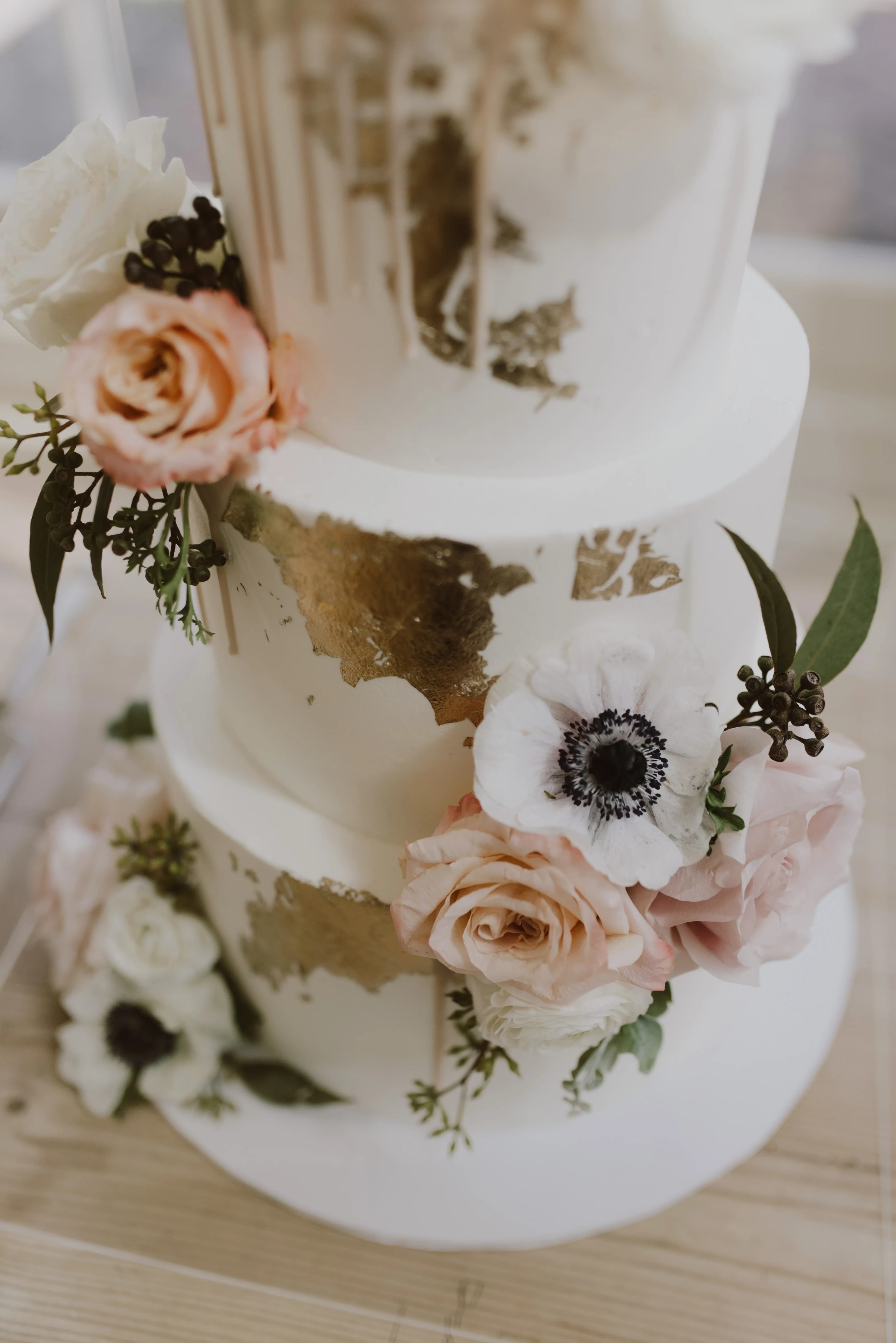 A three-tier white wedding cake decorated with gold leaf accents and adorned with white and peach roses, anemones, and greenery. The cake has a smooth texture with floral arrangements positioned around each tier, creating an elegant and romantic aesthetic.