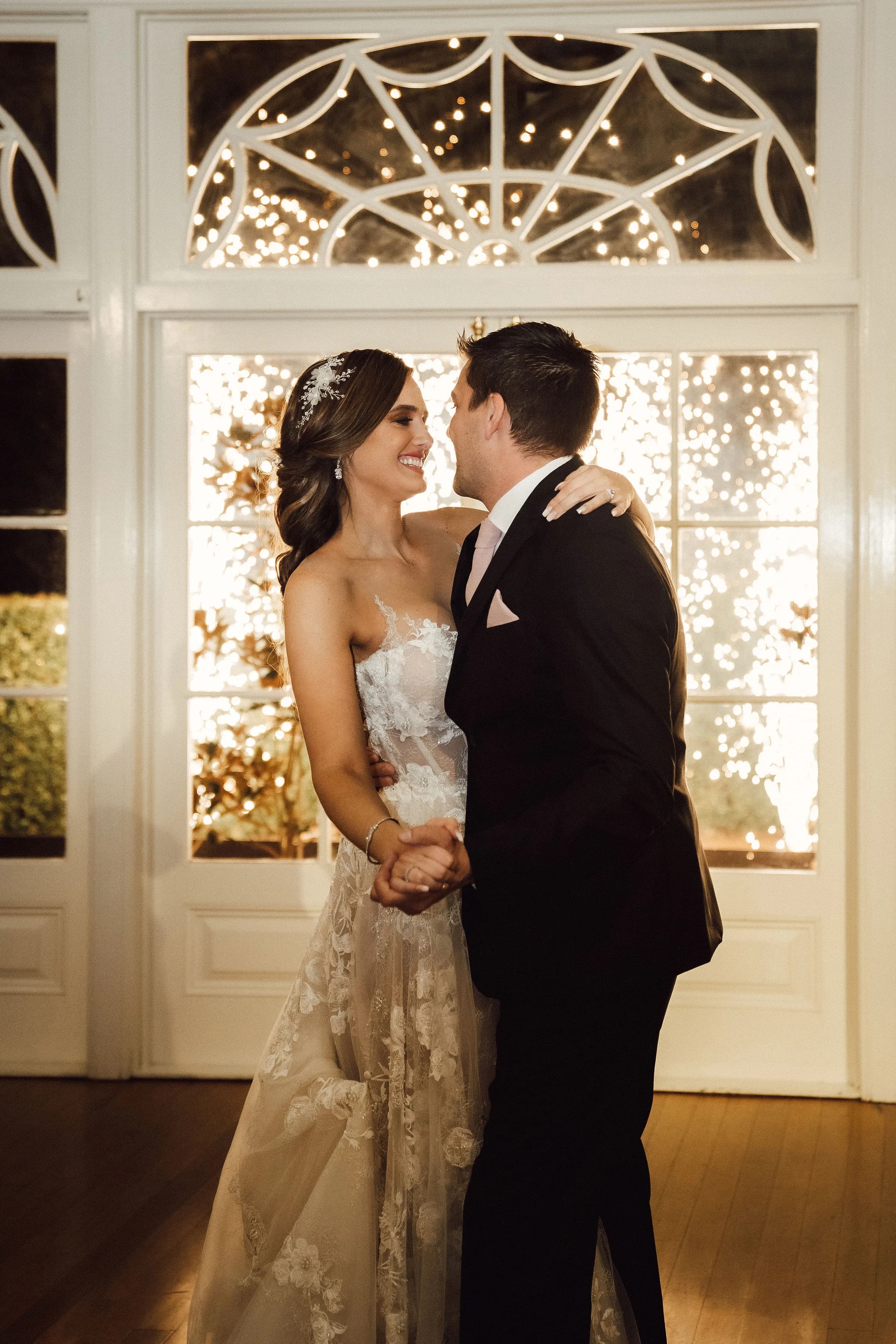 A bride and groom smile and embrace while dancing indoors. The bride wears a strapless, lace wedding gown, and the groom is in a black suit with a pink pocket square. Behind them, a large window is illuminated with twinkling lights, creating a festive atmosphere.