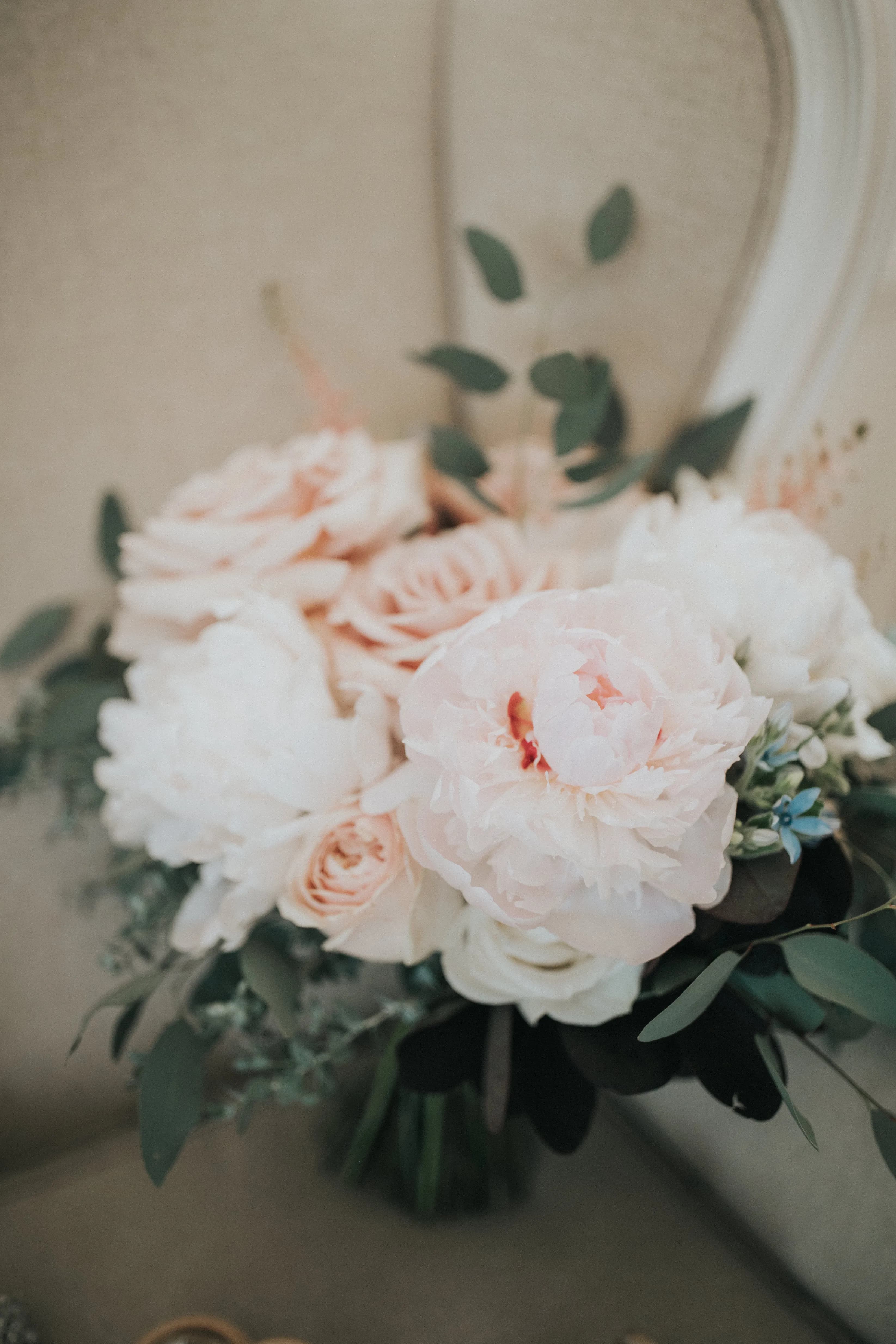 A floral bouquet featuring large, pale pink peonies and roses, accented with green foliage, rests on a light-colored, cushioned surface. The flowers appear fresh and delicately arranged.