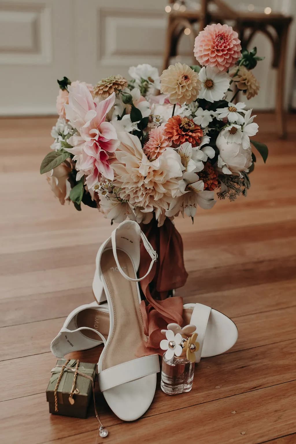 Bridal sandals, a bouquet of various flowers, a small wrapped gift, a perfume bottle, and a necklace with a pendant are elegantly arranged on a wooden floor.